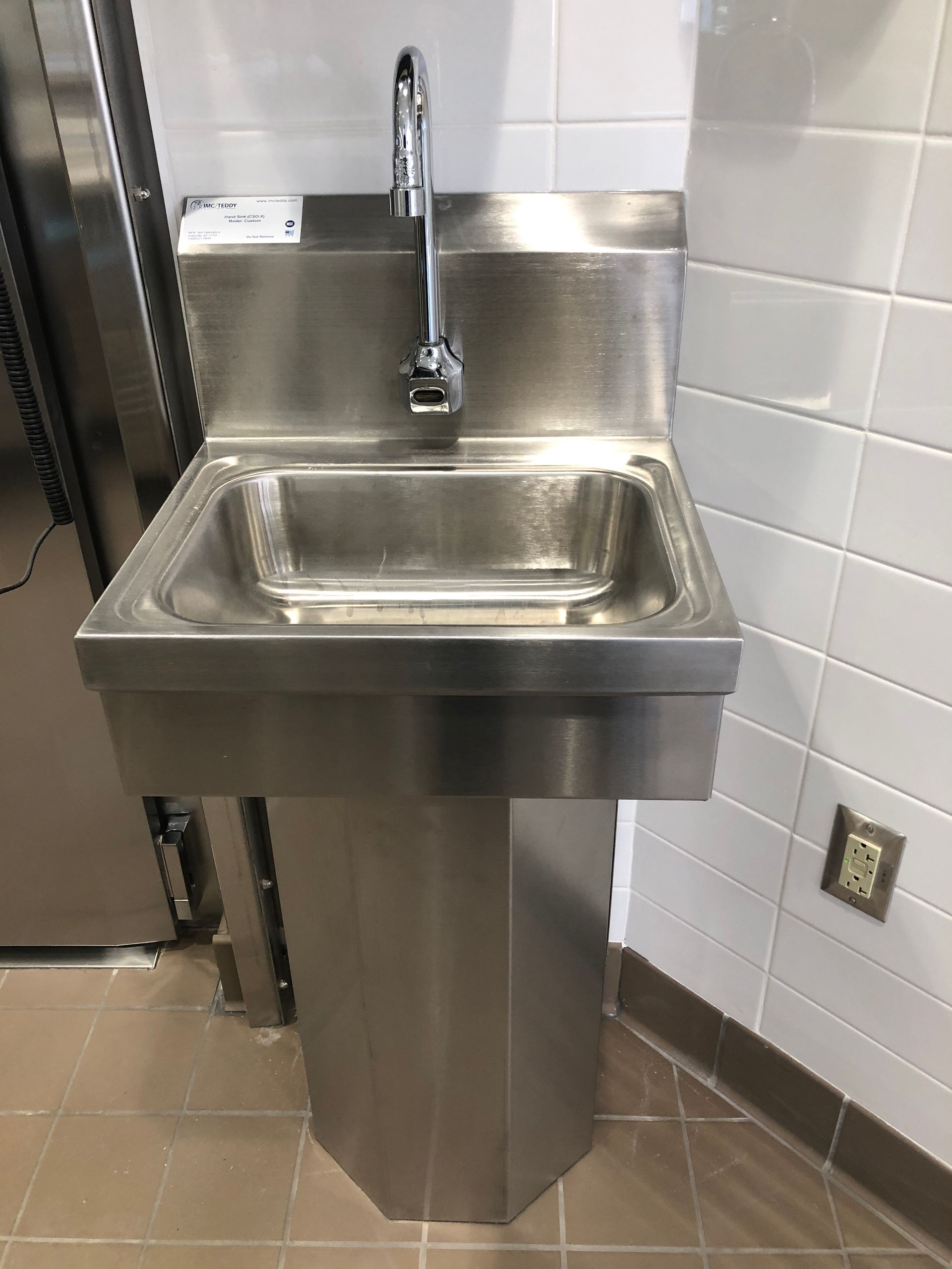 A stainless steel pedestal sink with a single faucet, mounted in a tiled room.
