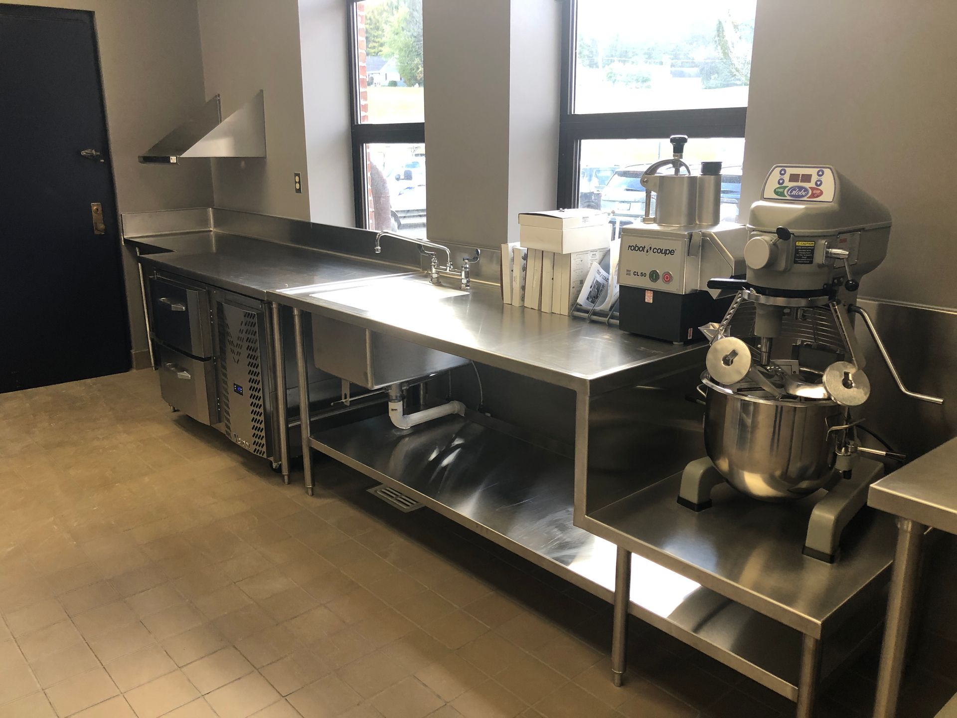 A stainless steel kitchen workspace featuring a commercial mixer, food processor, sink, and under-counter equipment.