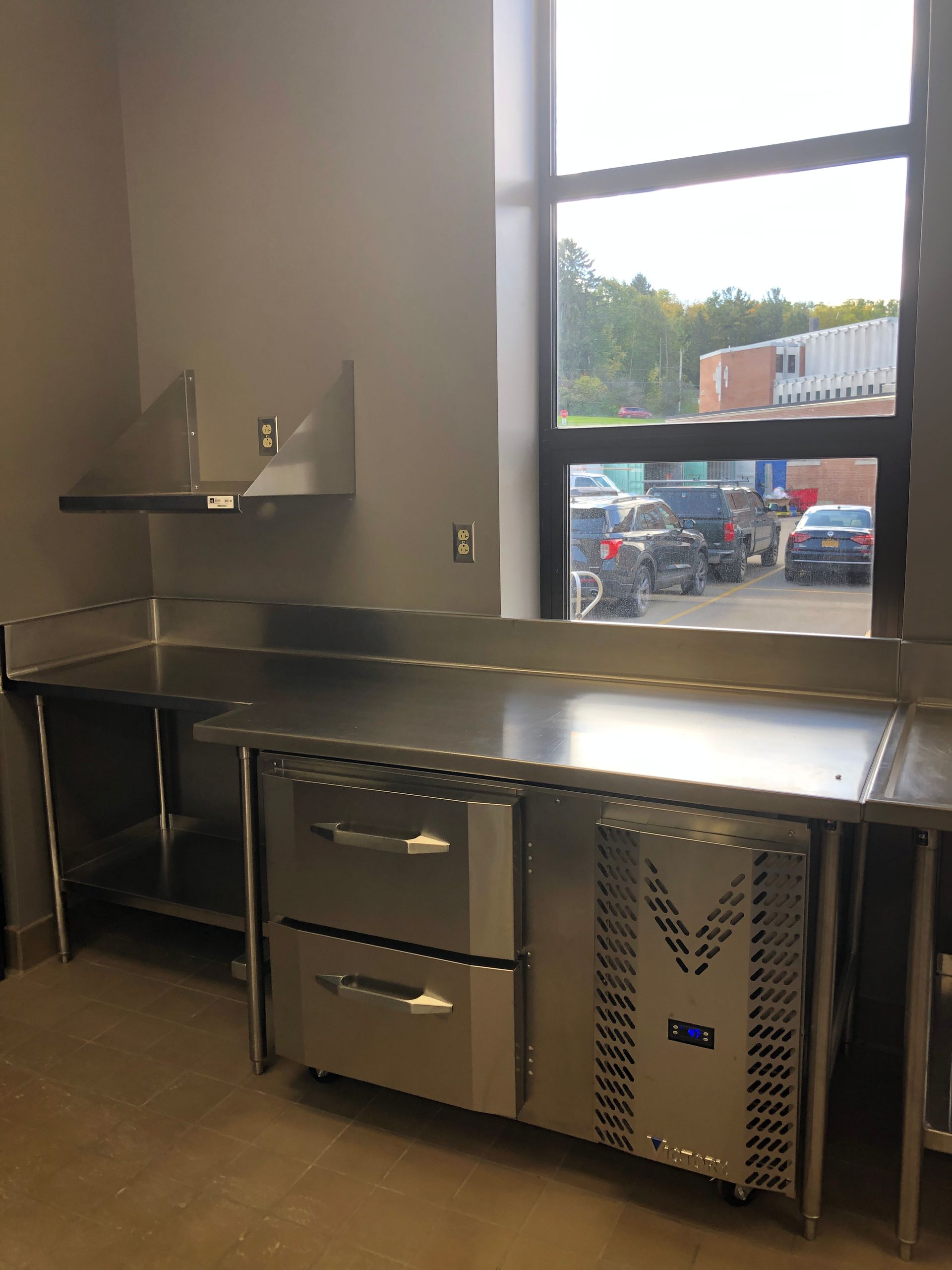 A stainless-steel commercial prep table with two built-in refrigerated drawers next to a large window.
