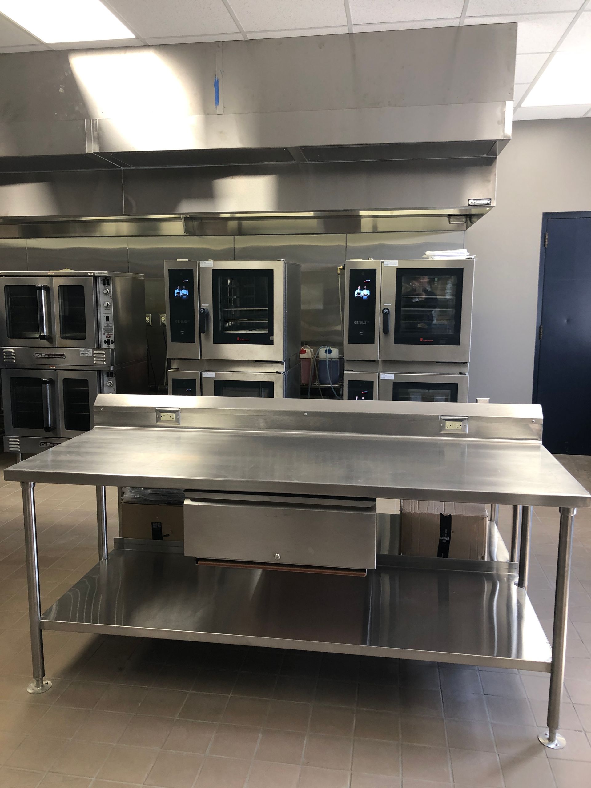 A stainless steel food prep table with a bottom shelf in a commercial kitchen featuring industrial ovens.