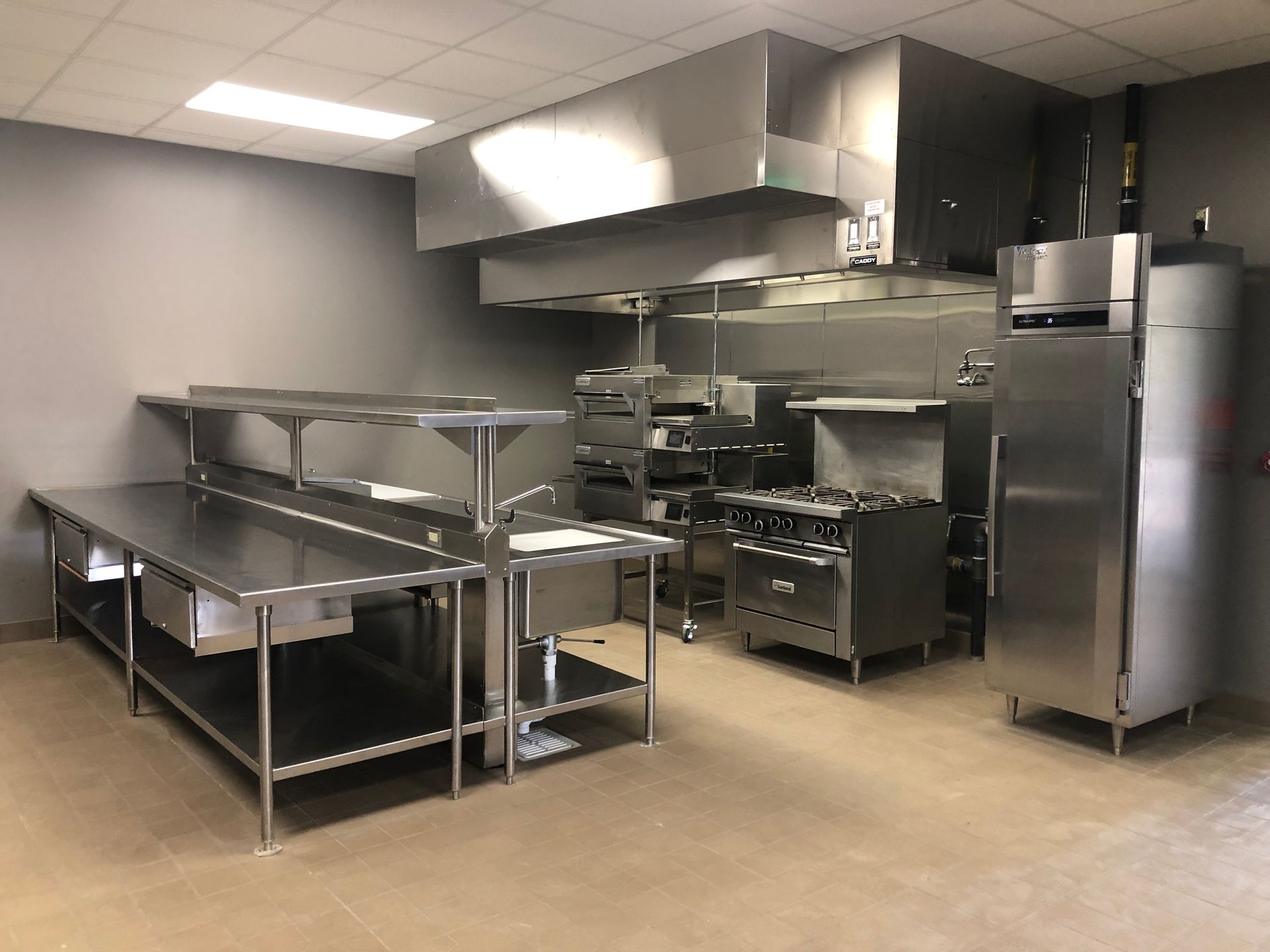 A modern commercial kitchen featuring stainless steel prep tables, a stove, an oven, and a tall refrigerator.