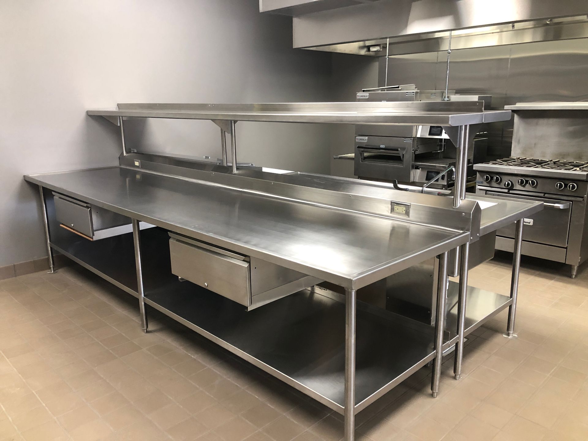 Commercial stainless steel food preparation table with an upper shelf and drawers in a professional kitchen.