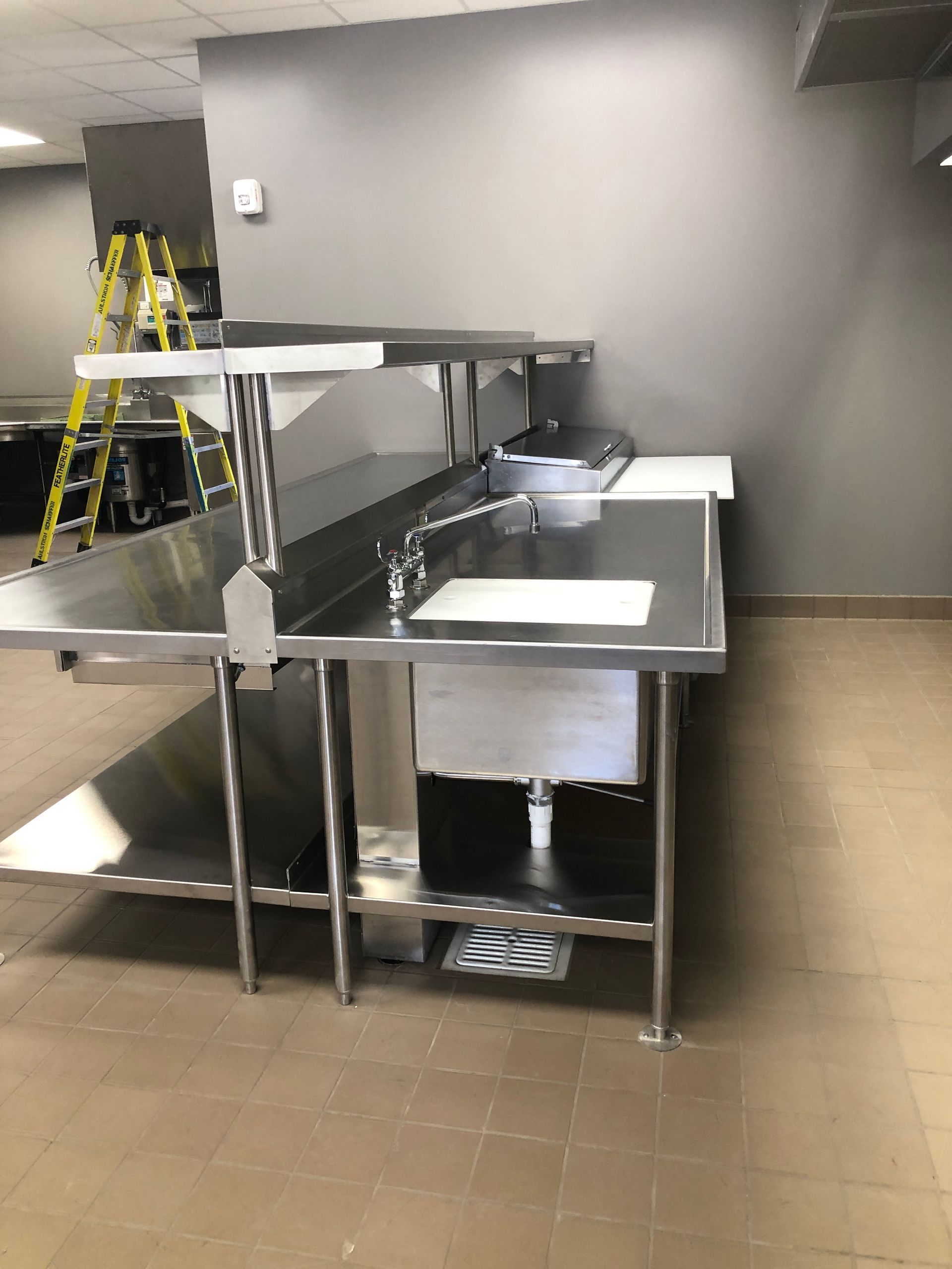 A stainless steel commercial sink with a shelving unit and faucet stands against a grey wall in a kitchen setting.