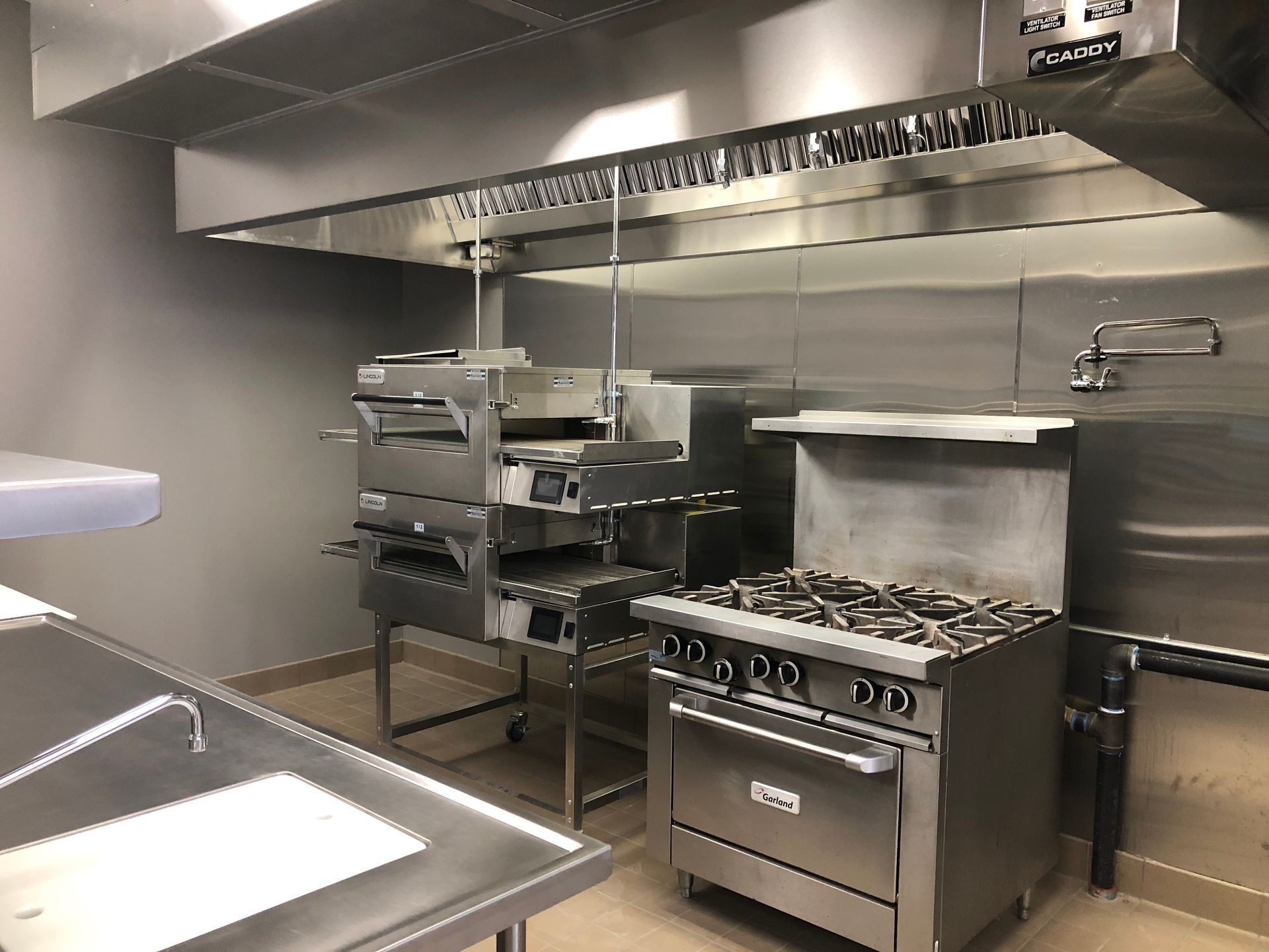A commercial kitchen featuring stainless steel pizza ovens, a gas range stove, and a sink beneath a metal ventilation hood.