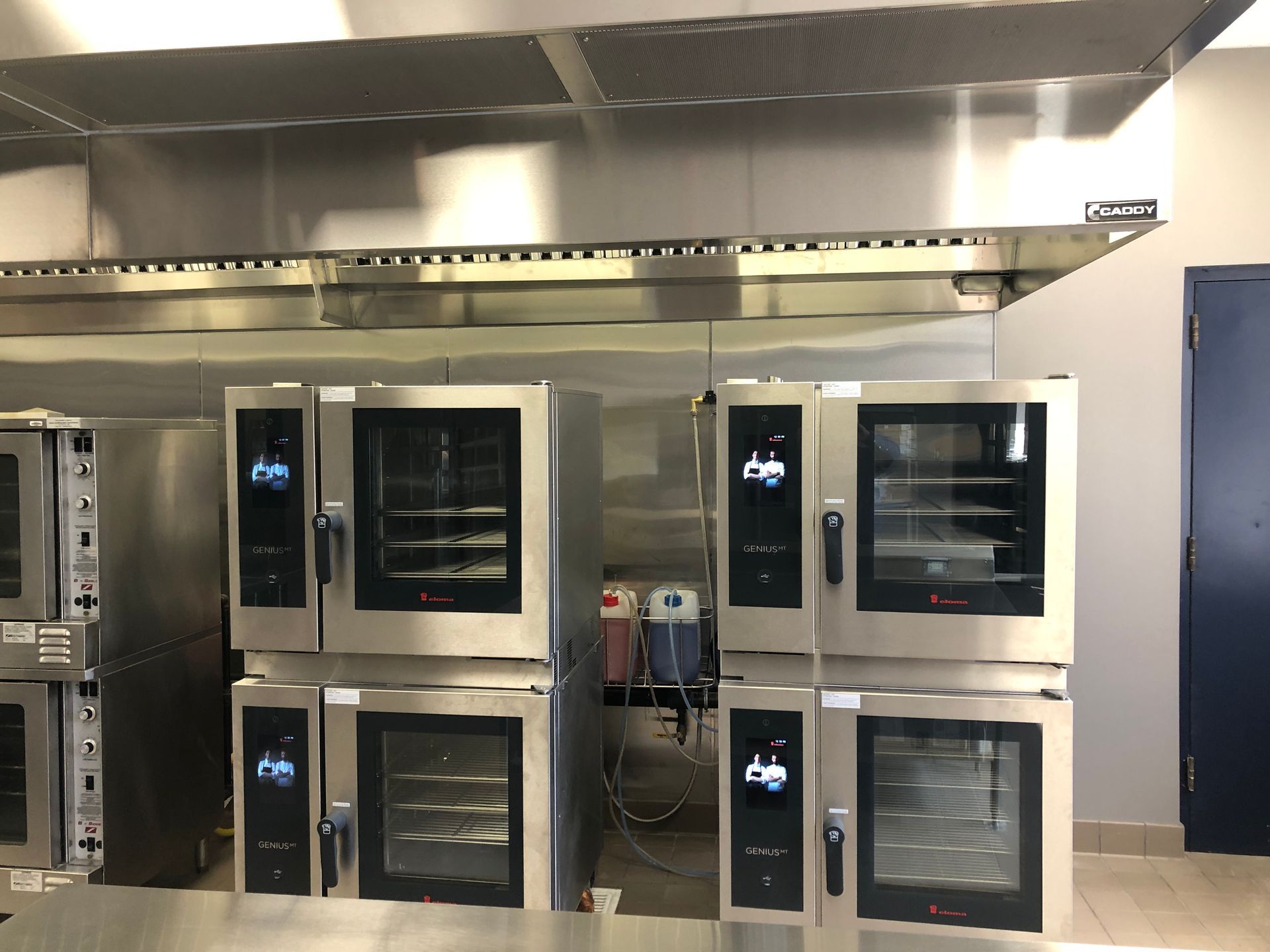 A commercial kitchen with stacked stainless steel convection ovens positioned under a large ventilation hood.