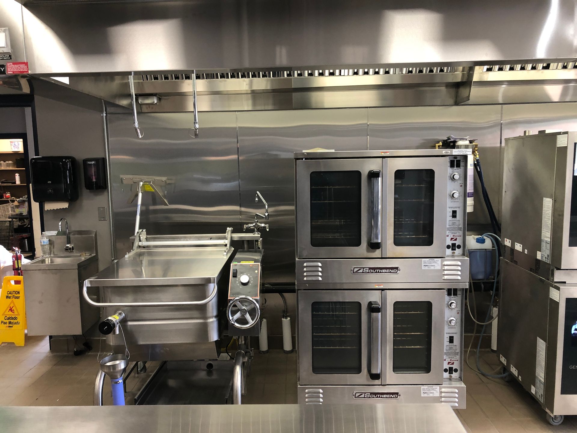 A professional kitchen features a tilting skillet, a stacked double oven, and a sink beneath a large stainless steel hood.