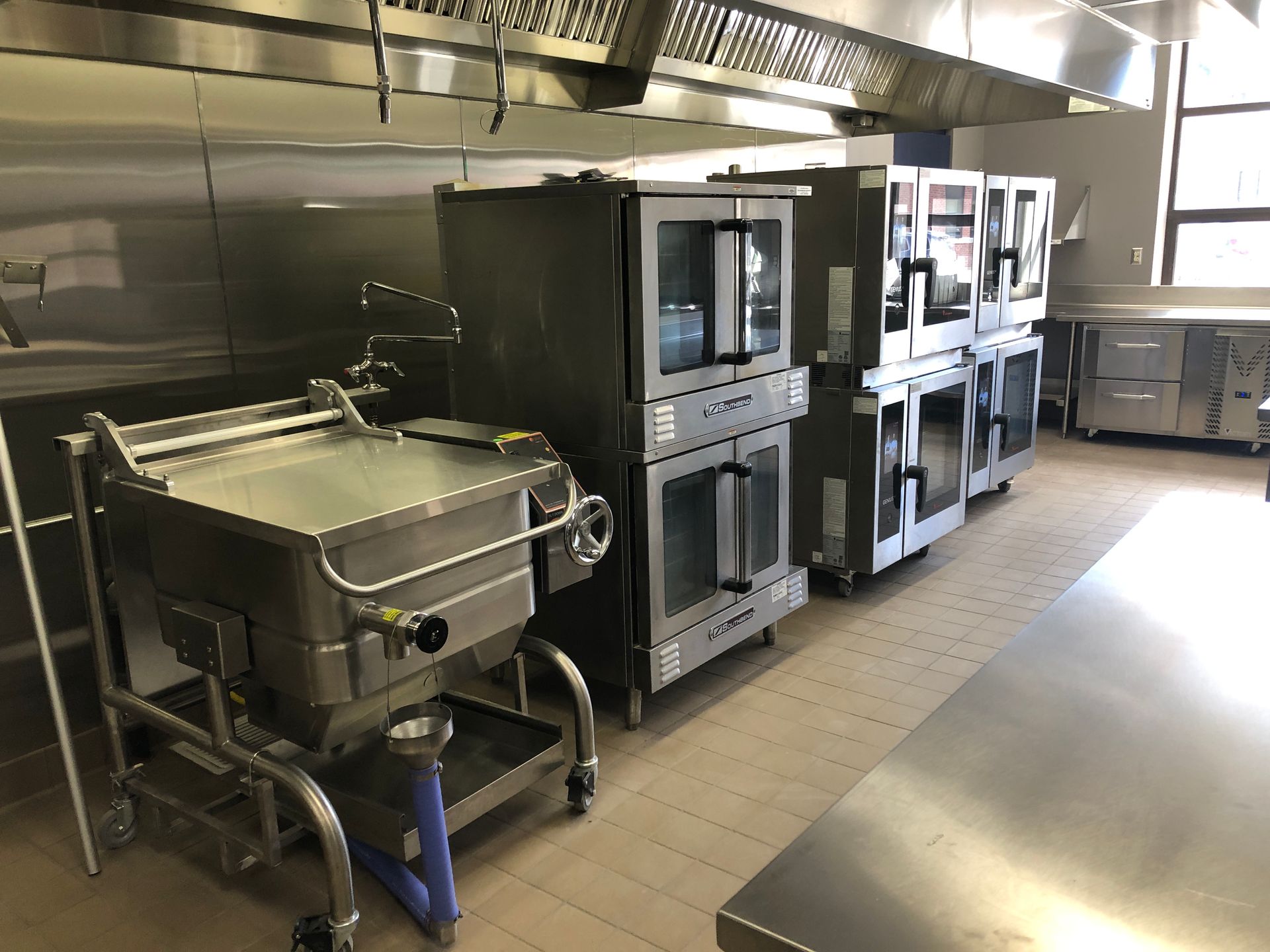 A stainless steel commercial kitchen featuring a tilting skillet and multiple stacked convection ovens under a vent hood.