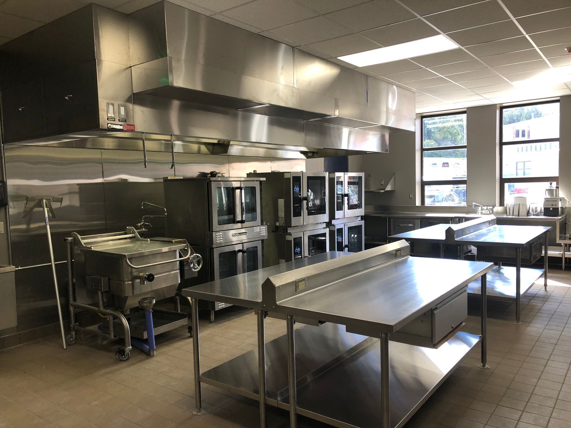 A clean, professional stainless steel commercial kitchen with prep tables, ovens, and a large ventilation hood.