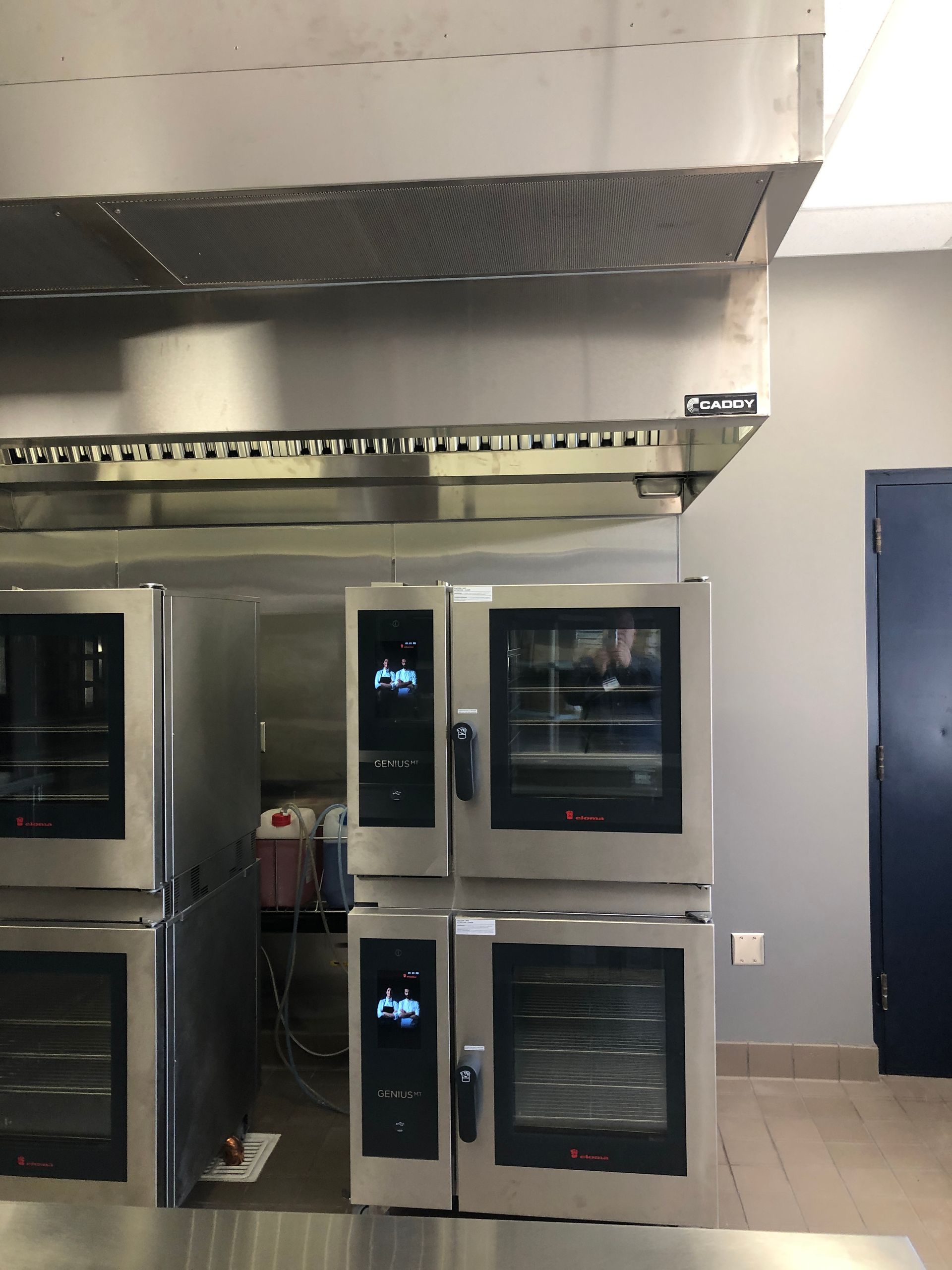 Stainless steel industrial stacked ovens sit under a vent hood in a professional kitchen.