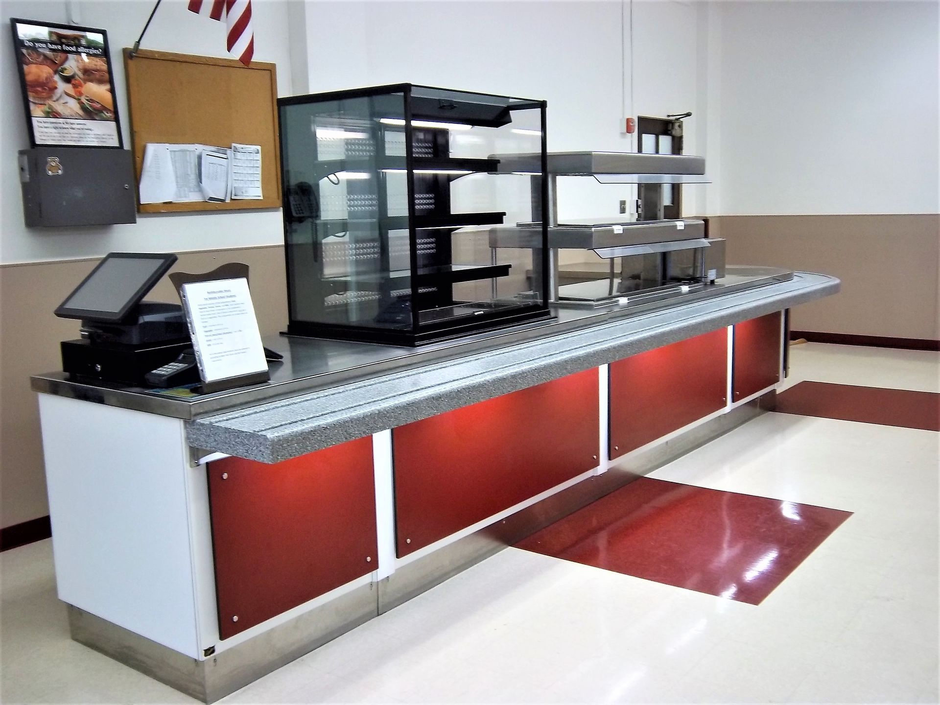 A food service counter featuring a glass display case, a point-of-sale system, and red panels in a brightly lit room.