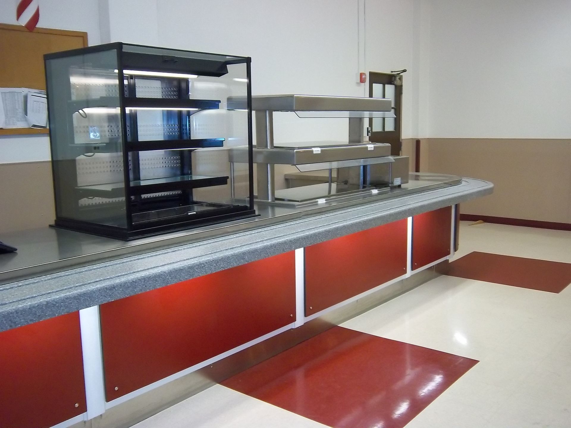 A serving counter in a kitchen with a glass display case and metal racks, featuring red panels on a light-colored floor.