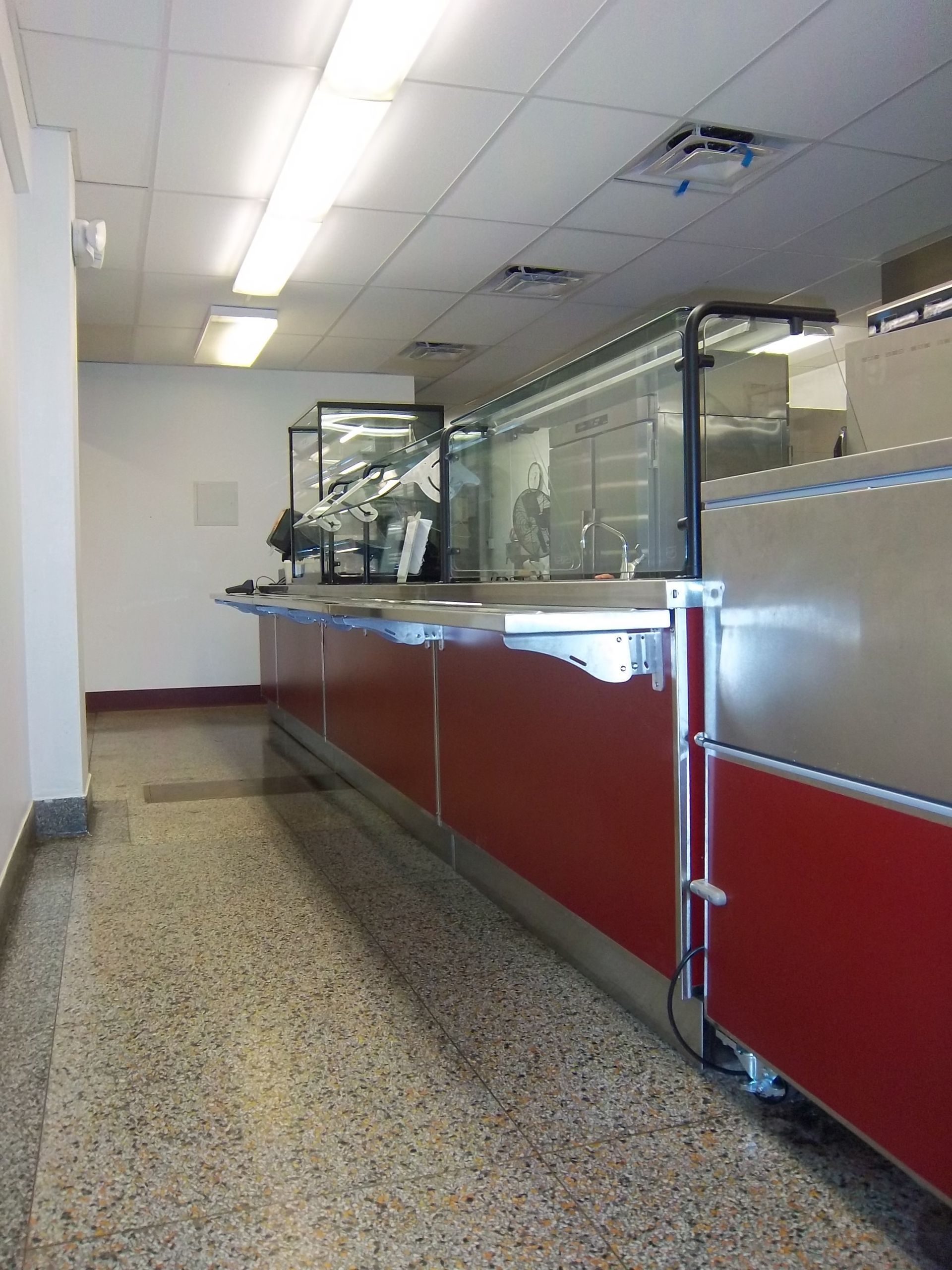A perspective view of a long, red-paneled service counter with a glass sneeze guard, set in a room with speckled flooring.