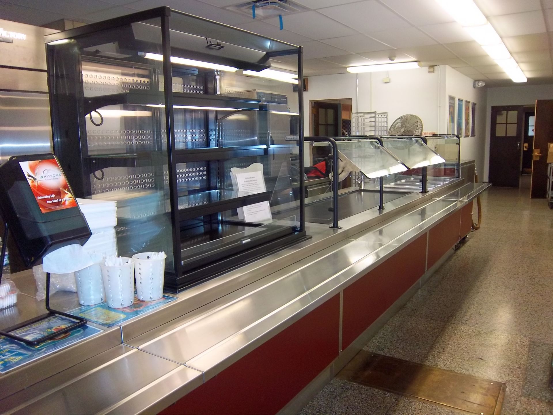 A stainless steel food service counter with a tall glass display case and serving trays in a brightly lit dining area.