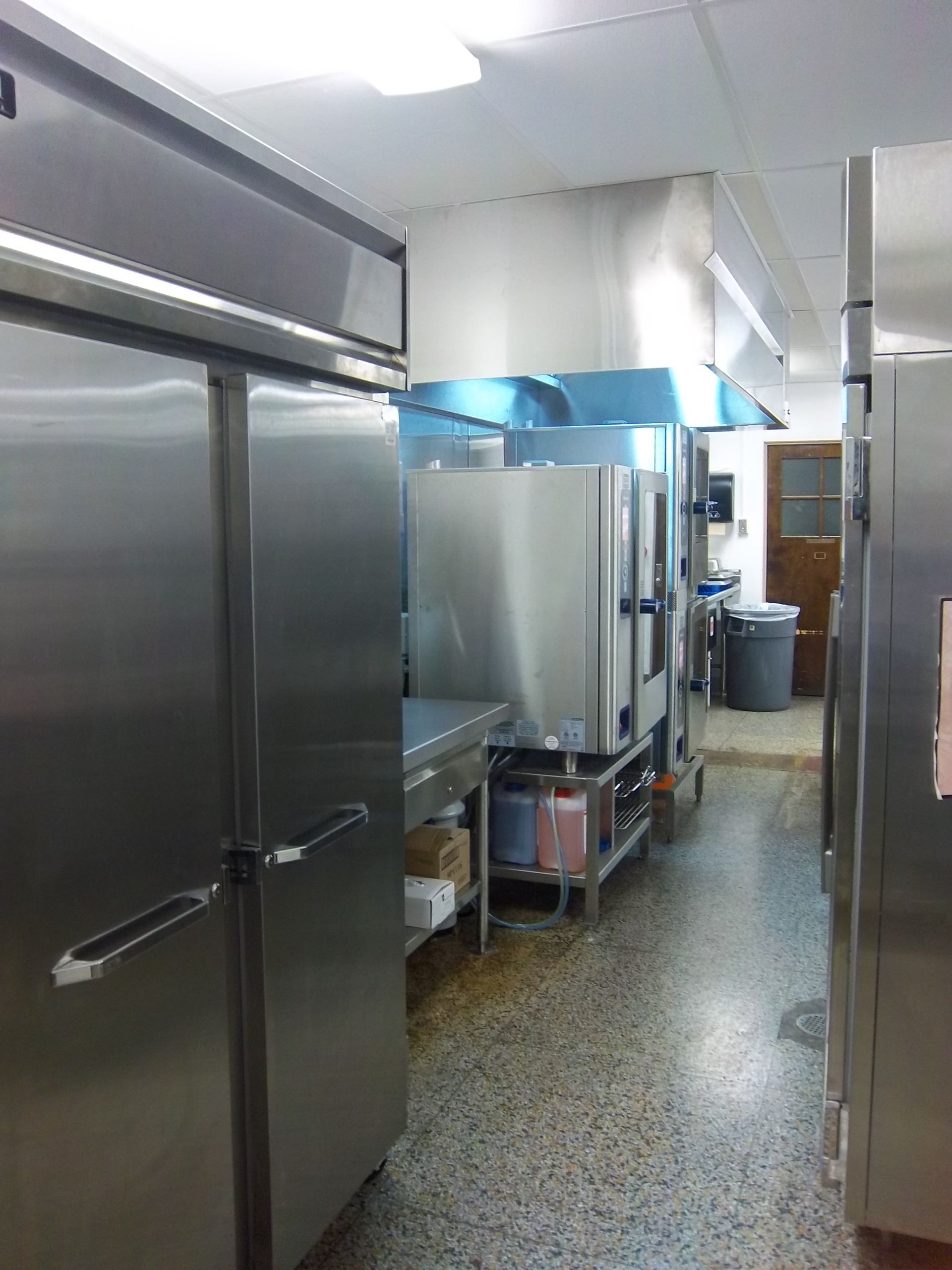 A commercial kitchen with large stainless steel refrigerators and ovens under an industrial vent hood on a speckled floor.