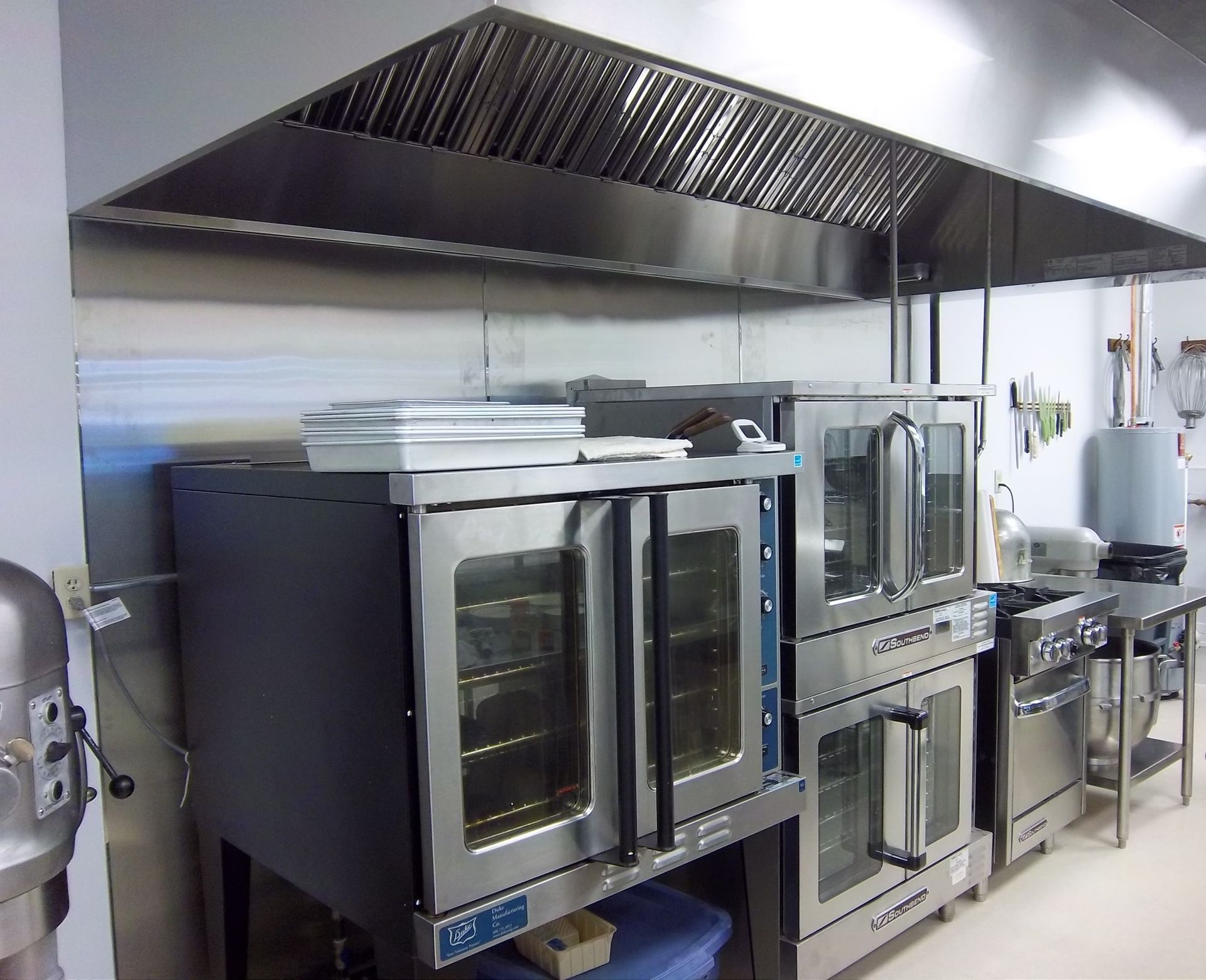 Stainless steel commercial kitchen with convection ovens and a large ventilation hood over the cooking area.