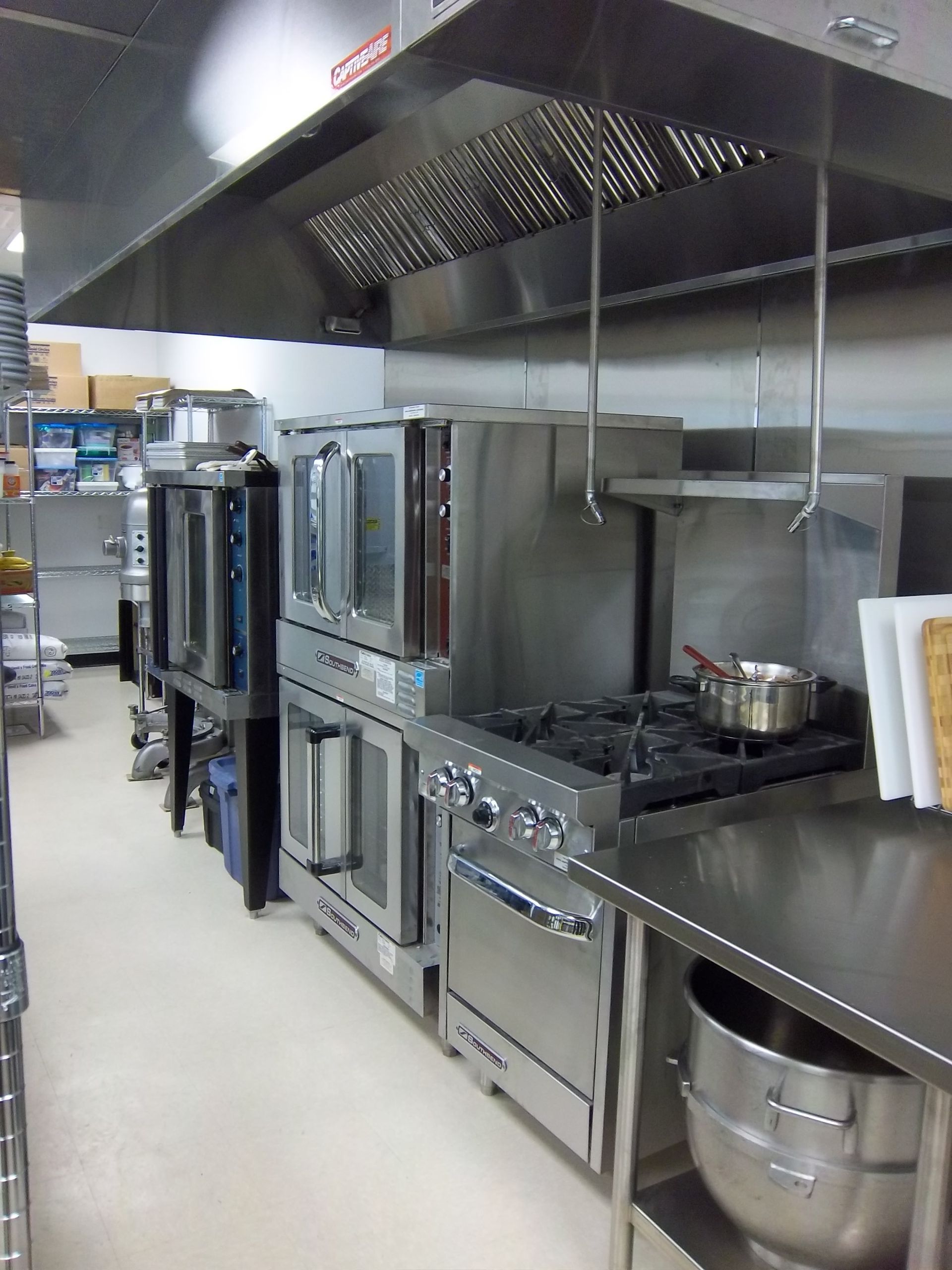 A stainless steel commercial kitchen line with ovens, a stovetop, and a large mixing bowl on a prep table.