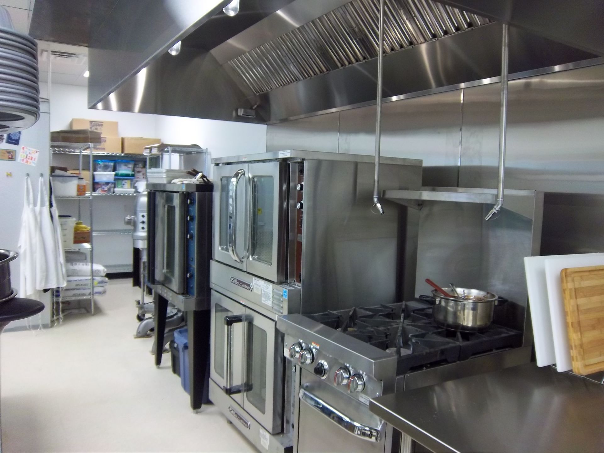 A commercial kitchen featuring stainless steel stoves, ovens, and a vent hood above, with storage shelves in the background.