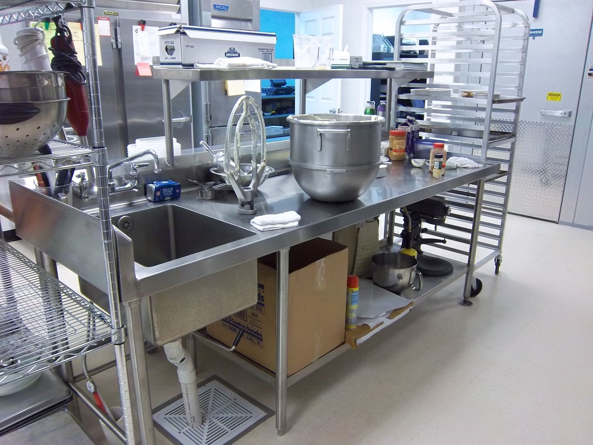 A stainless steel kitchen prep station with a sink, large mixing bowl, and storage rack in a commercial kitchen setting.