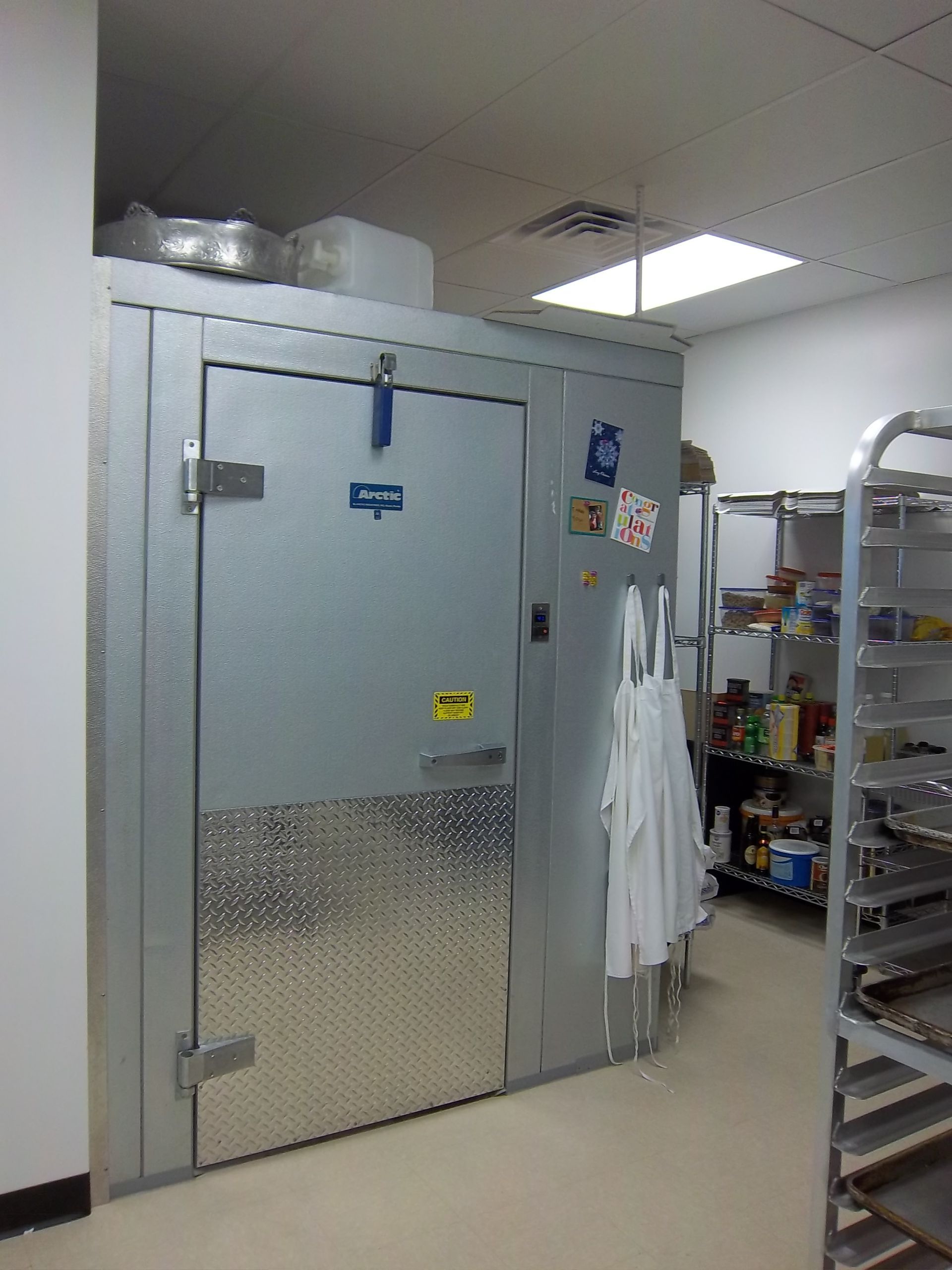 A large metal walk-in cooler door in a kitchen, with white aprons hanging on the side and pantry shelves in the background.