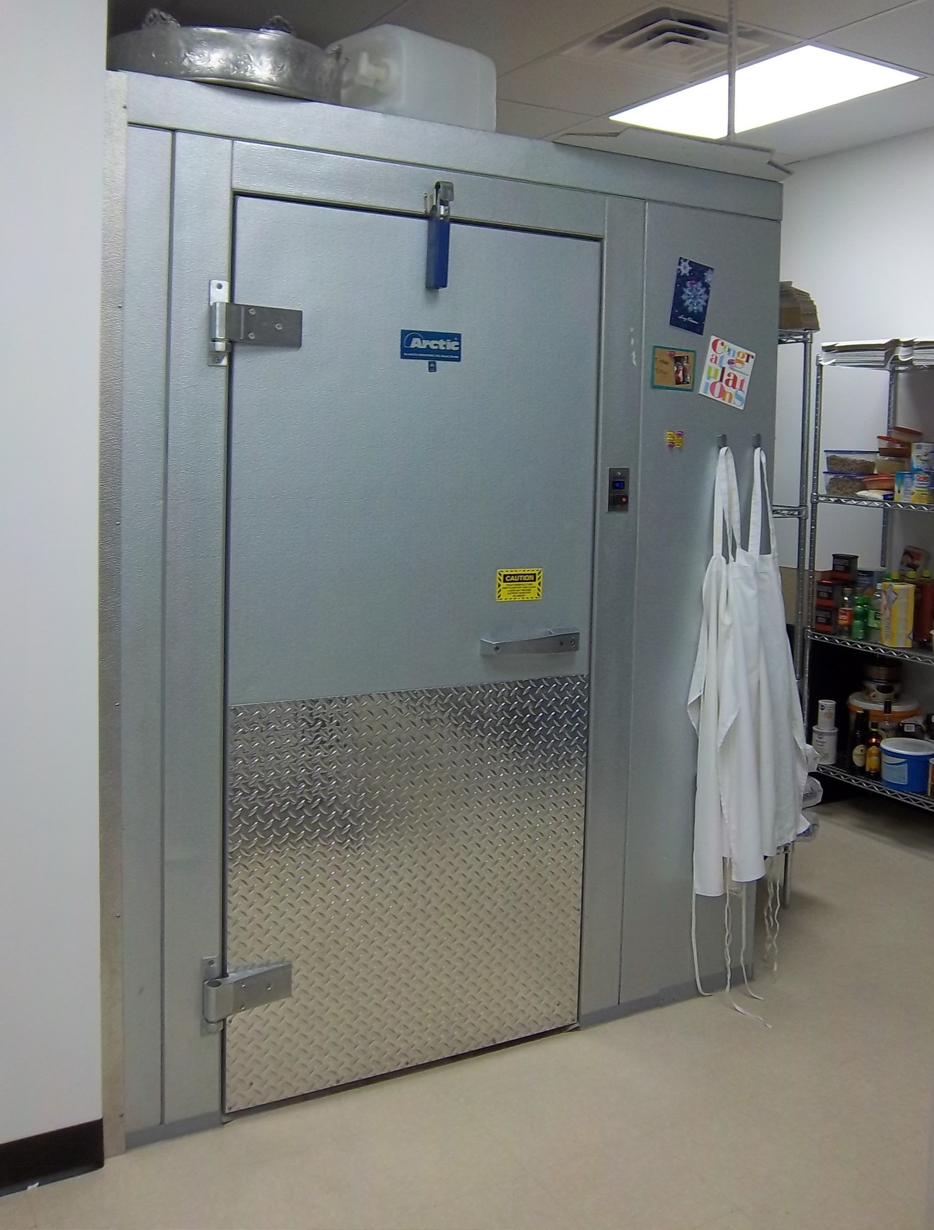 A commercial walk-in freezer door with diamond-plate steel cladding on the bottom, located in a storage area.