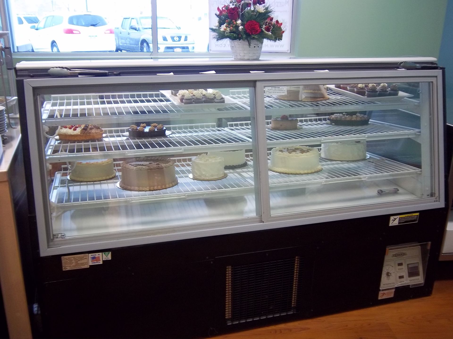 A refrigerated bakery display case containing multiple cakes on wire shelves, with a small flower arrangement on top.