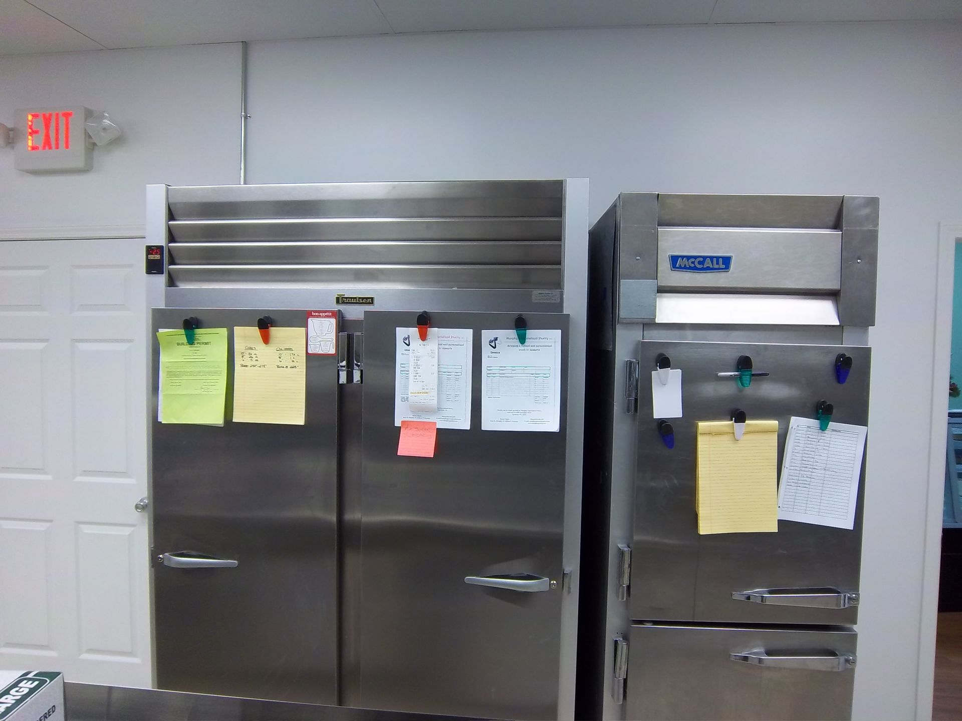 Two commercial stainless-steel refrigerators stand side-by-side against a white wall, with papers attached to their doors.