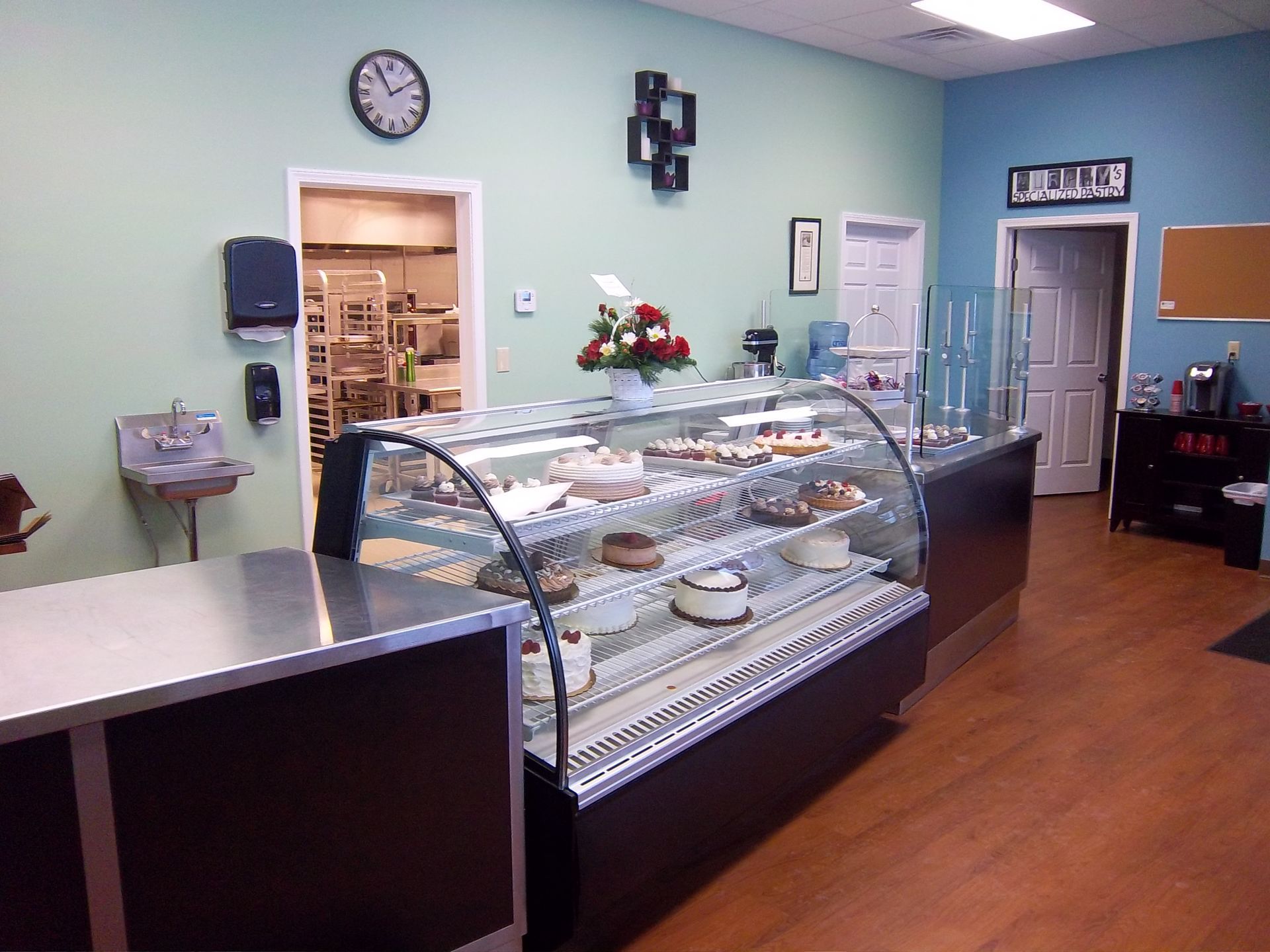A bakery interior featuring a glass display case filled with cakes, light green and blue walls, and a wood-look floor.