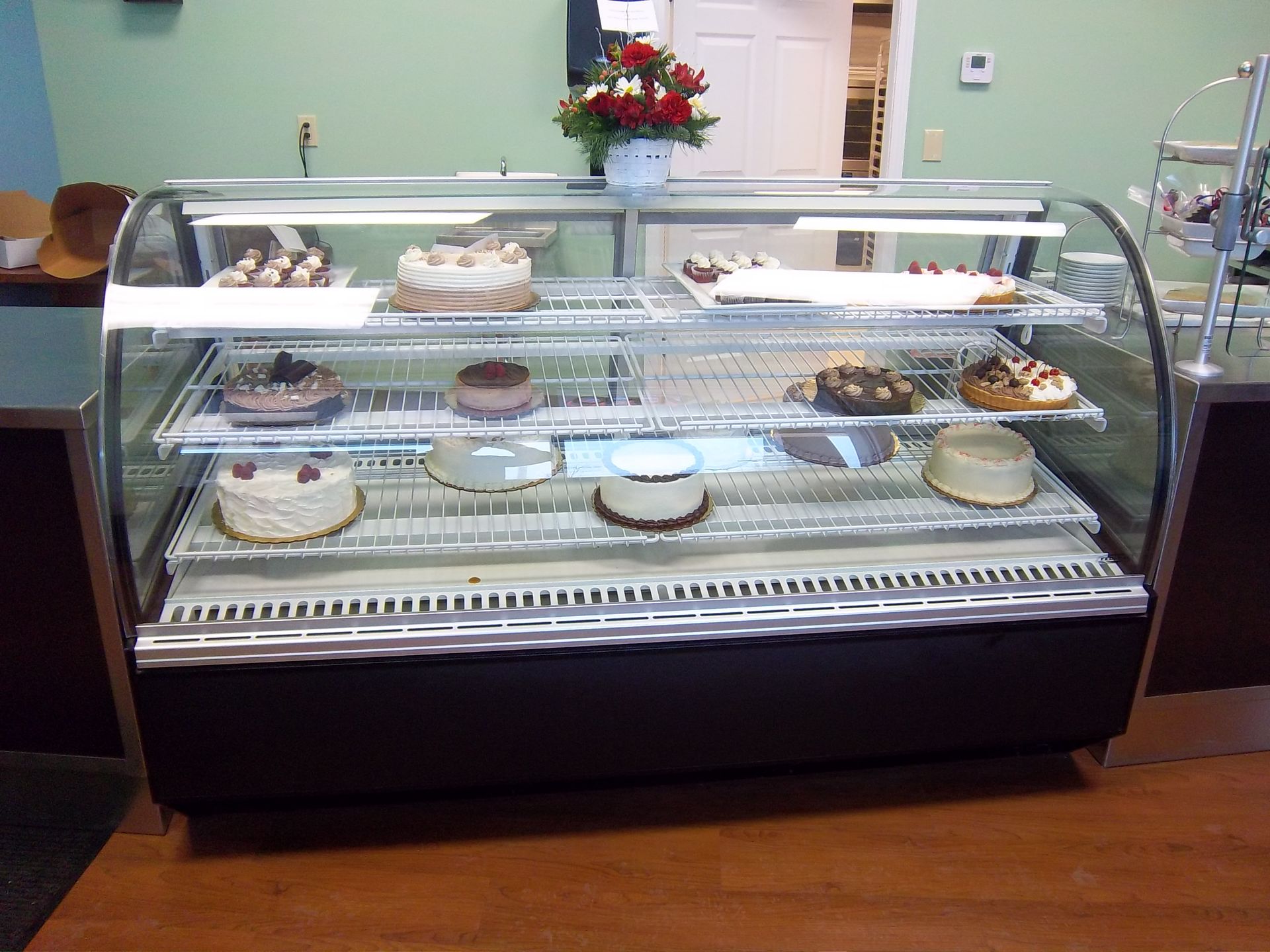 A refrigerated bakery display case holds various cakes and small desserts on three shelves, topped with flower decor.