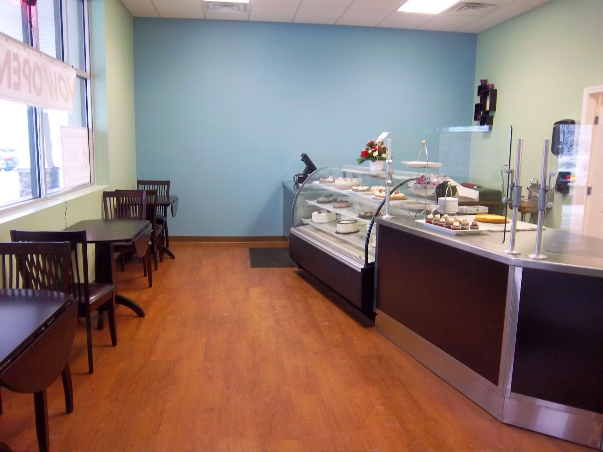 A bakery interior with light blue walls, a glass display case, and small tables with chairs on a wood-look floor.