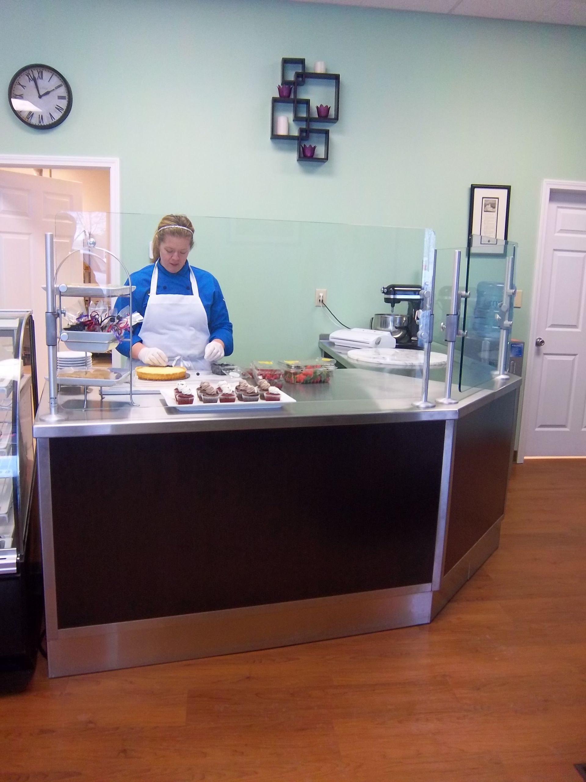 A person in a blue shirt and white apron prepares pastries behind a stainless steel counter in a bakery.