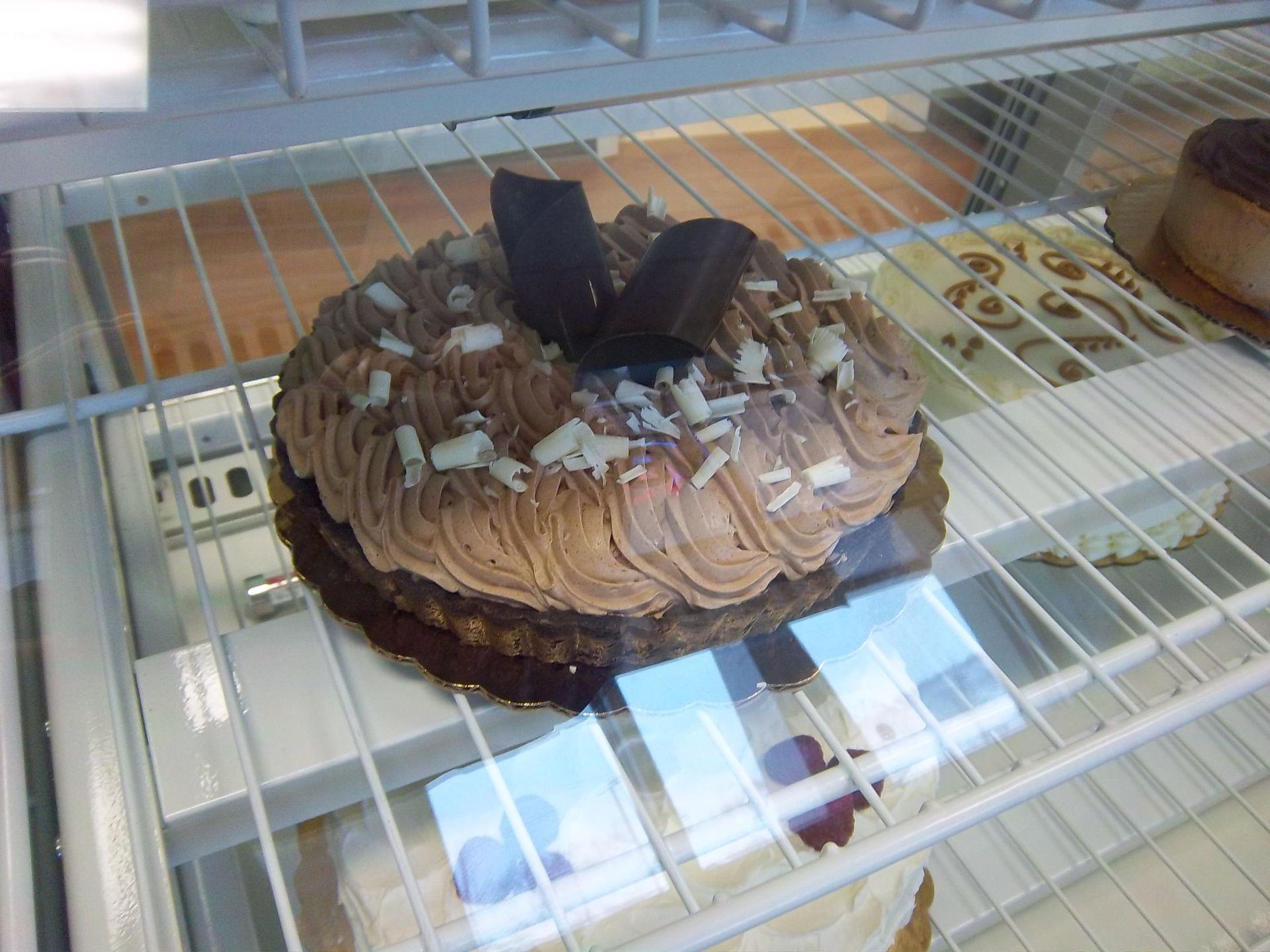 A chocolate cake with piped frosting and white chocolate shavings, displayed on a wire shelf in a glass bakery case.