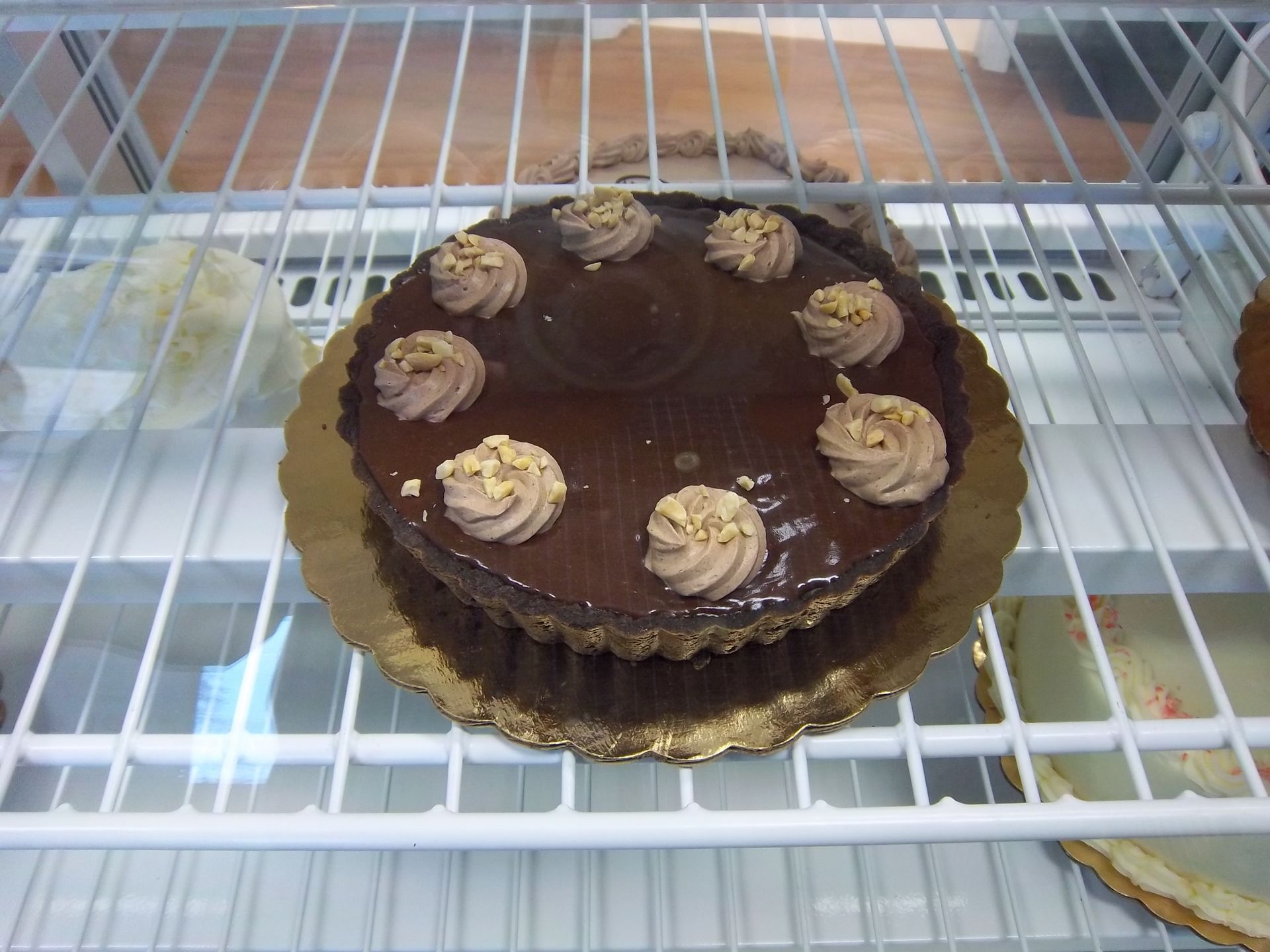 A chocolate tart topped with eight piped buttercream swirls and a sprinkle of nuts, sitting on a rack in a refrigerated case.