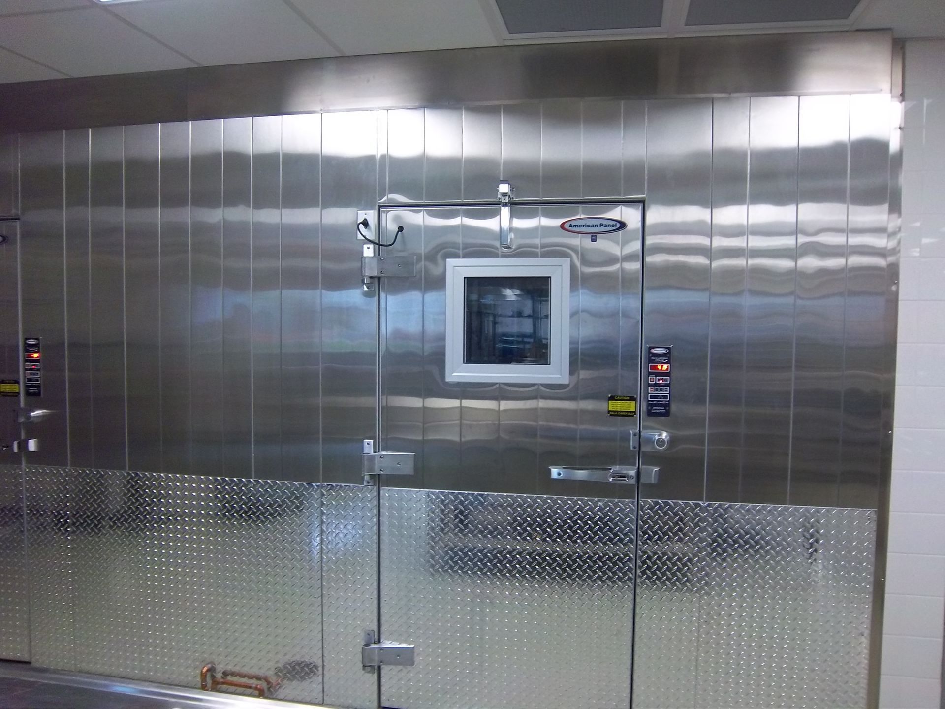 A walk-in commercial stainless steel refrigerator or freezer with a diamond-plate lower panel and a windowed door.