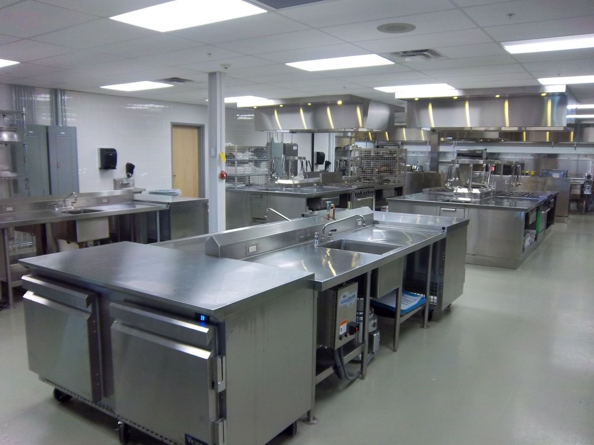 A spacious professional kitchen with rows of stainless steel work tables, prep sinks, and overhead ventilation hoods.