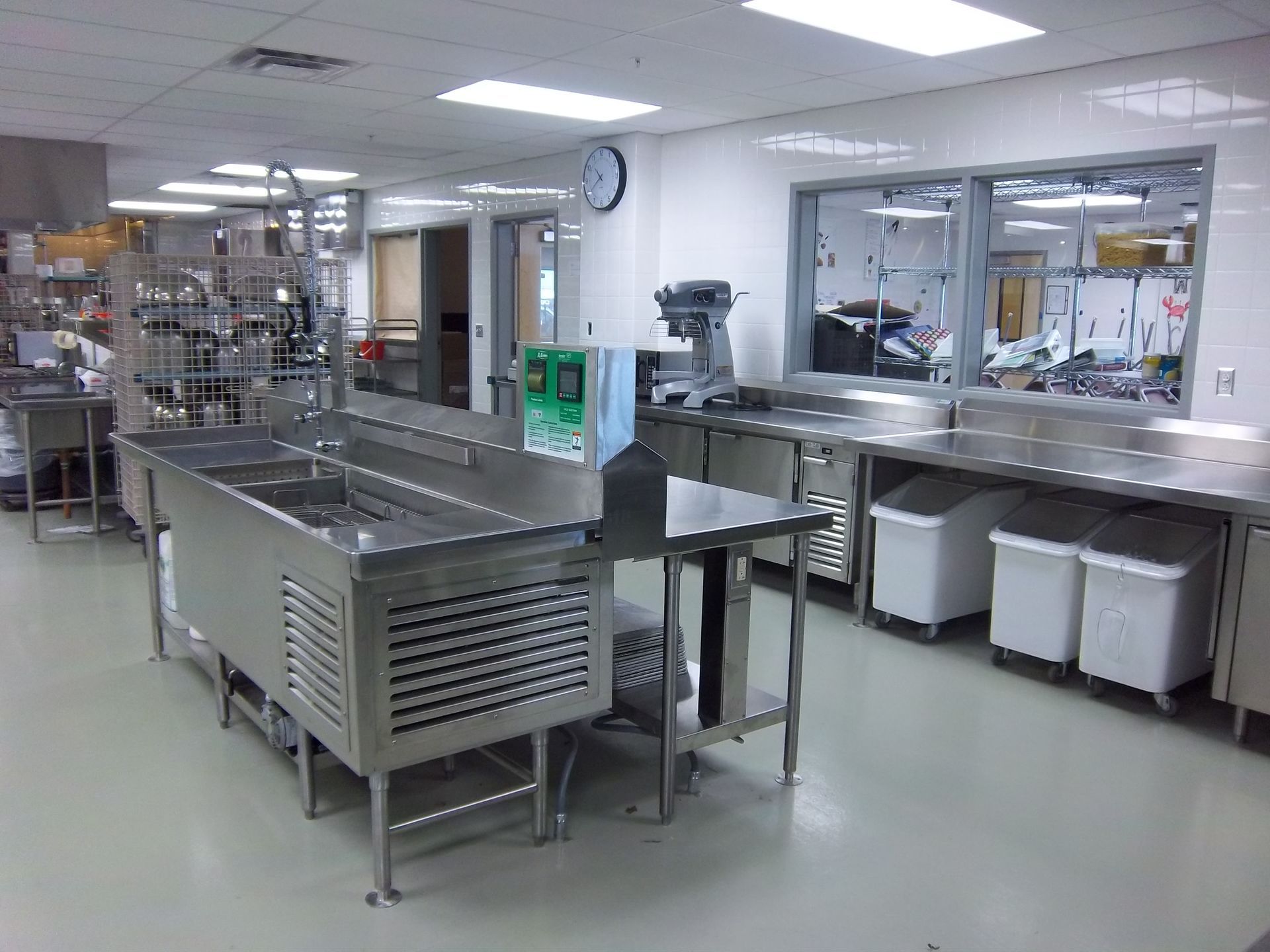 A commercial kitchen featuring stainless steel prep tables, a sink, a standing mixer, and white ingredient bins.