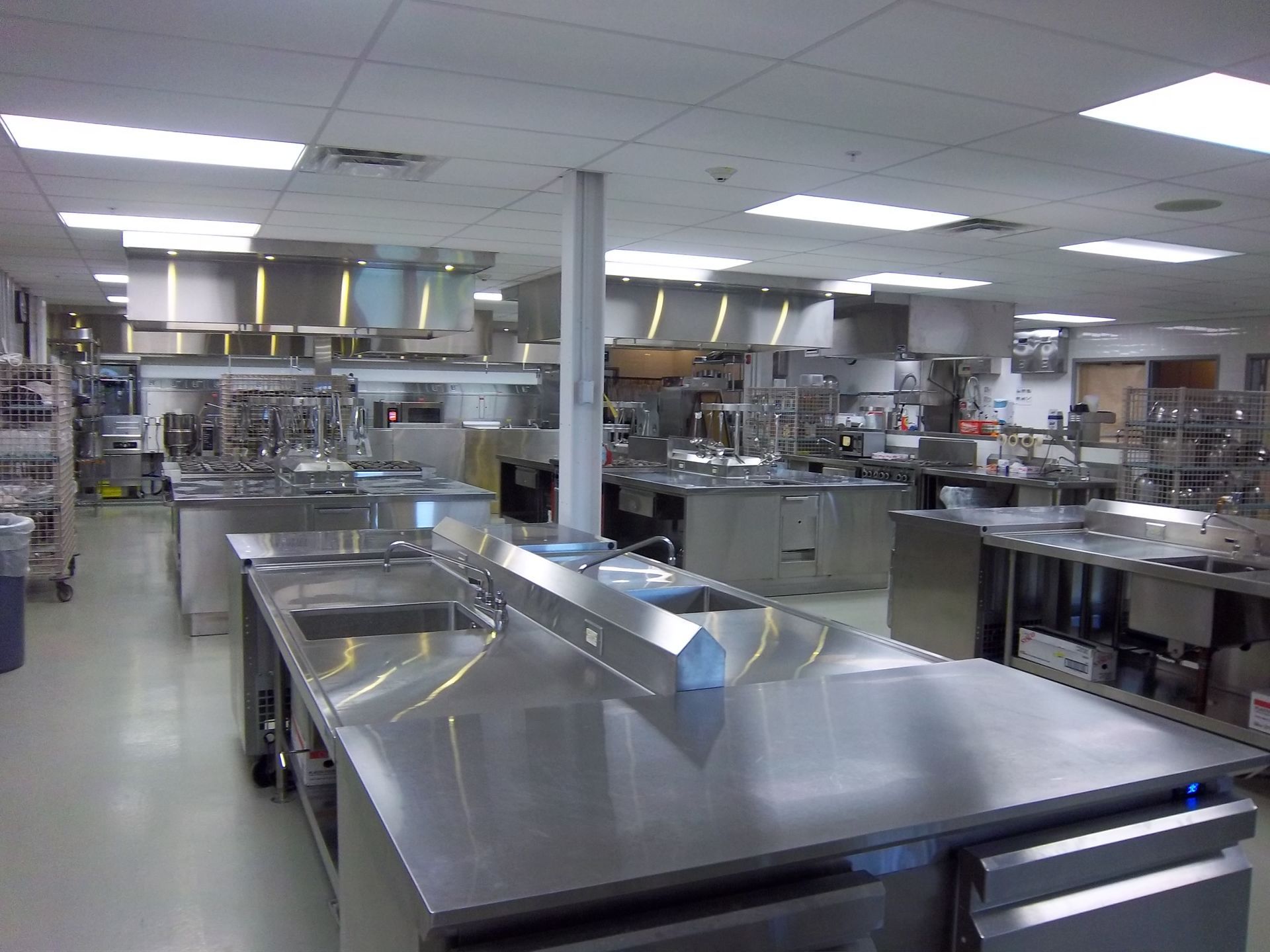A professional stainless steel commercial kitchen with multiple workstations, sinks, and overhead ventilation hoods.