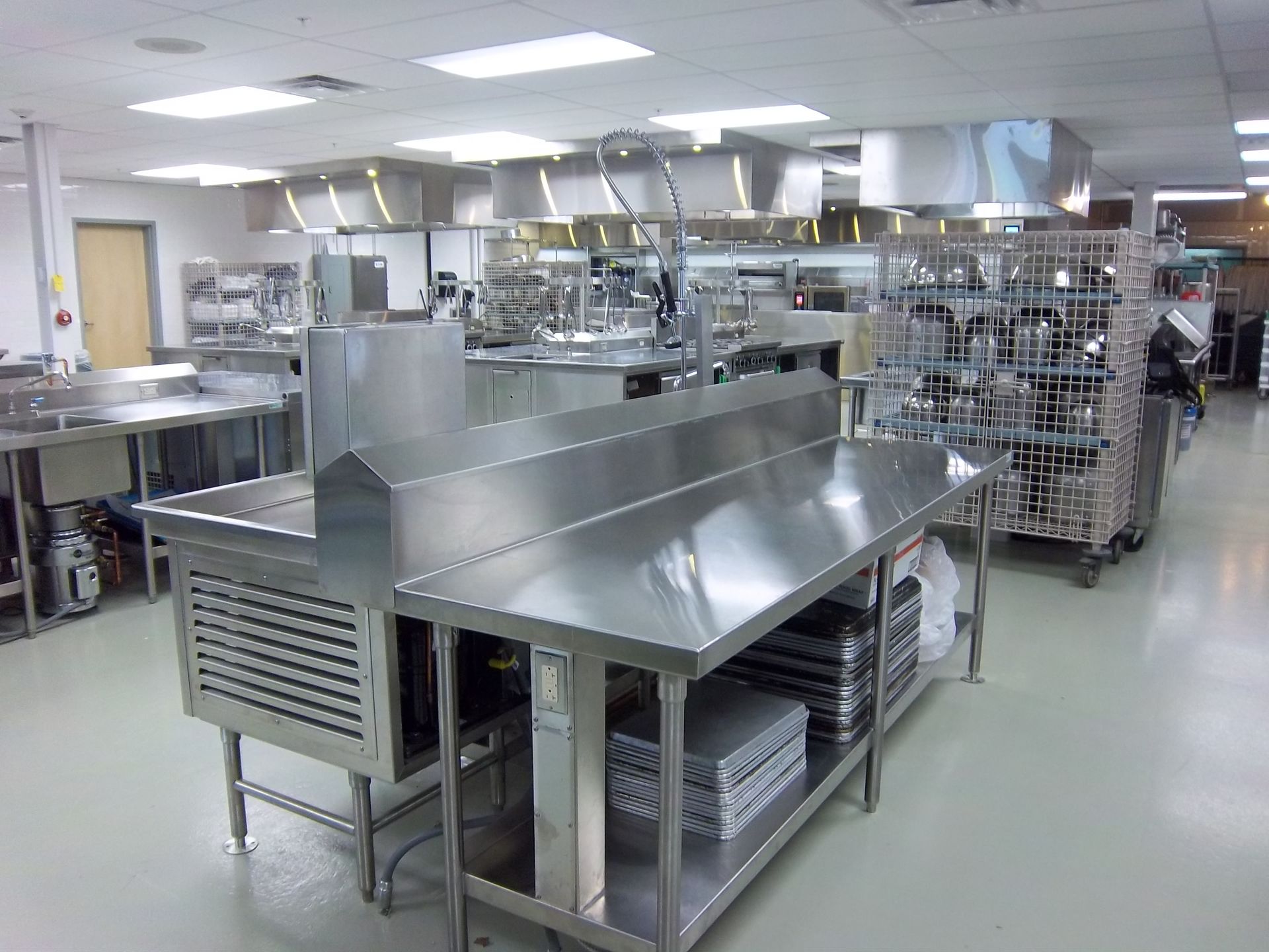Commercial kitchen with stainless steel prep tables, refrigerated units, and metal shelving racks on a light-colored floor.