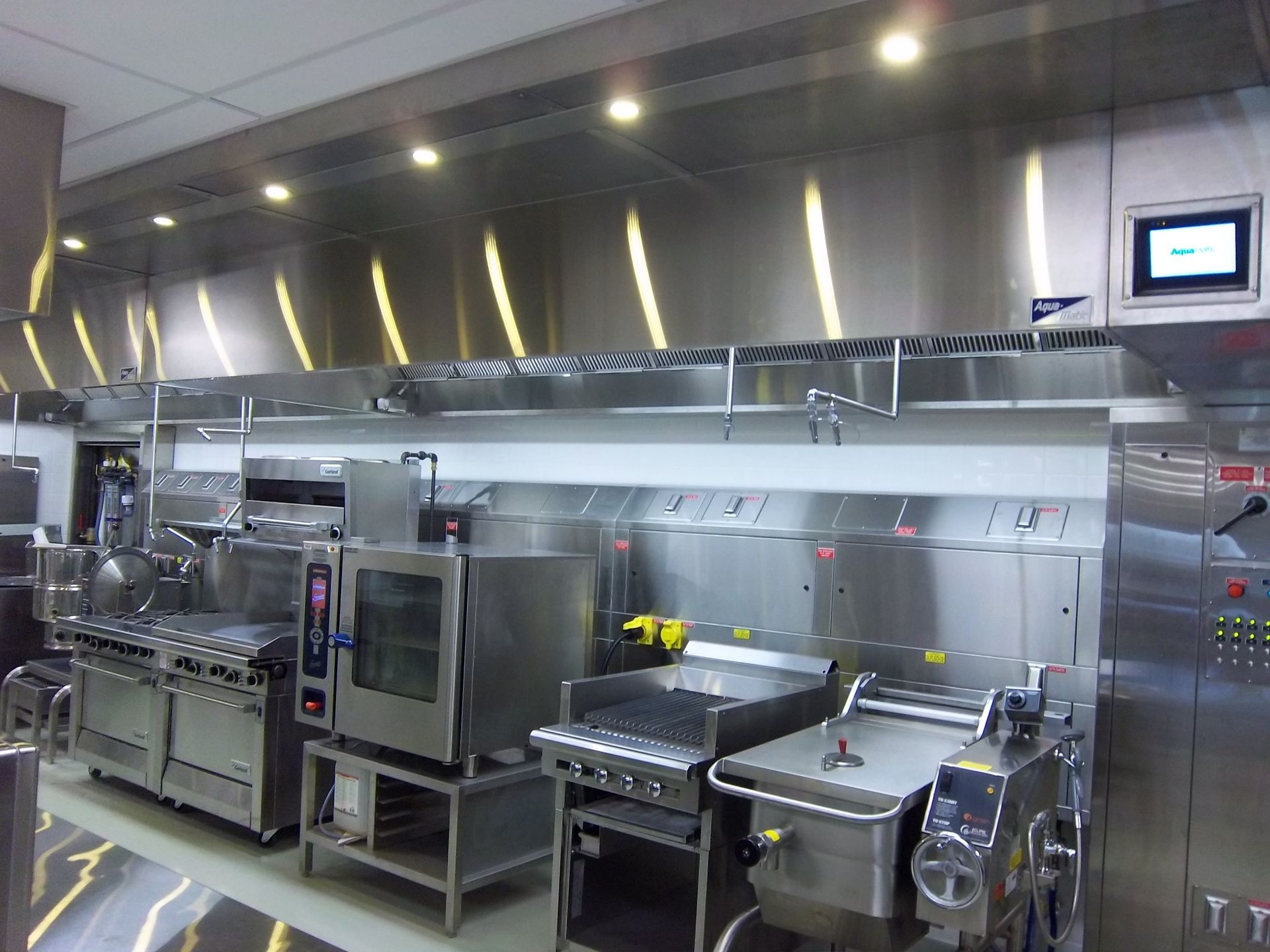 A stainless steel professional kitchen line featuring an oven, range, flat-top grill, and tilt skillet under a vent hood.