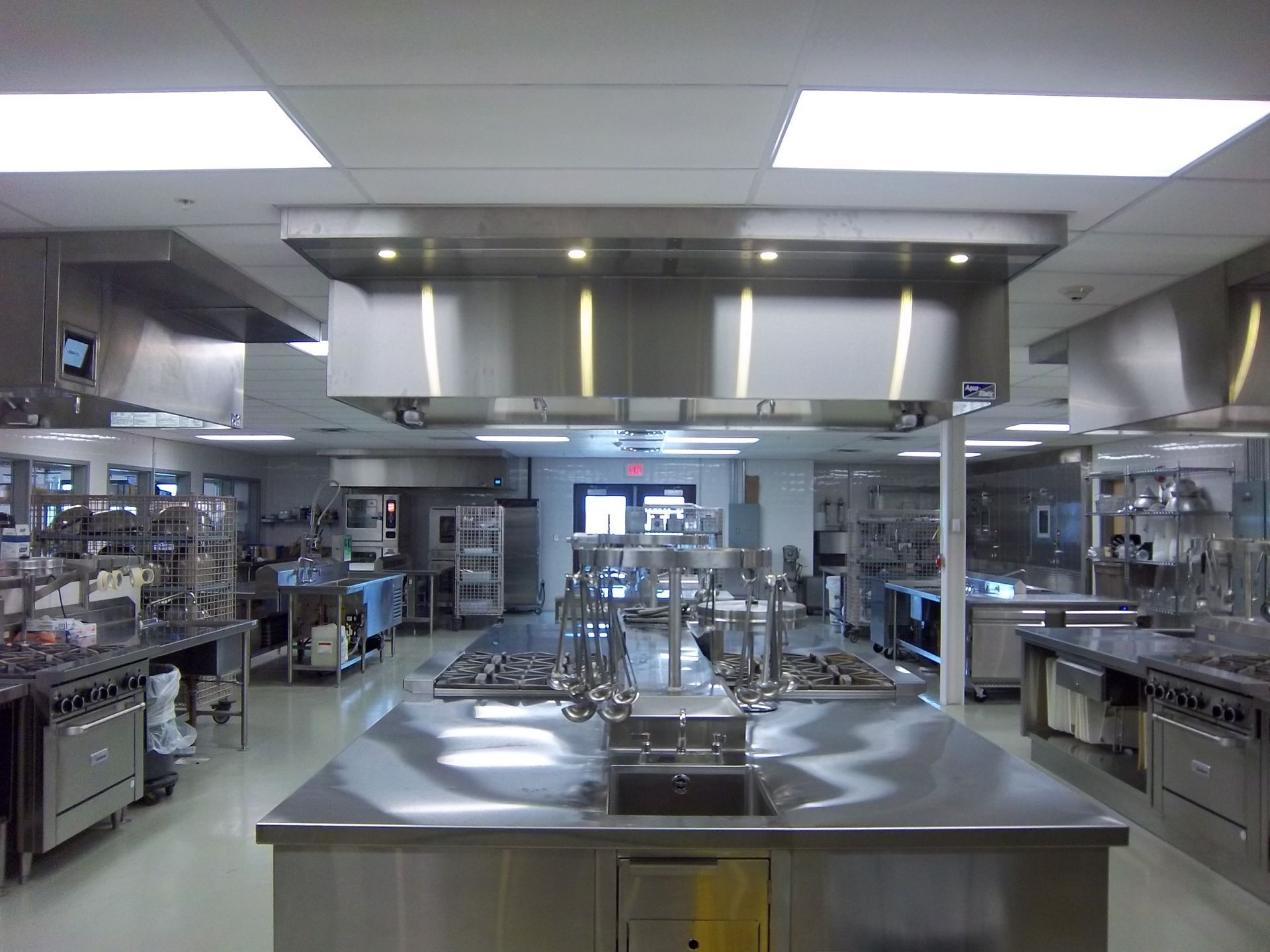 A well-lit, professional kitchen workspace with stainless steel counters, cooking ranges, and overhead ventilation hoods.