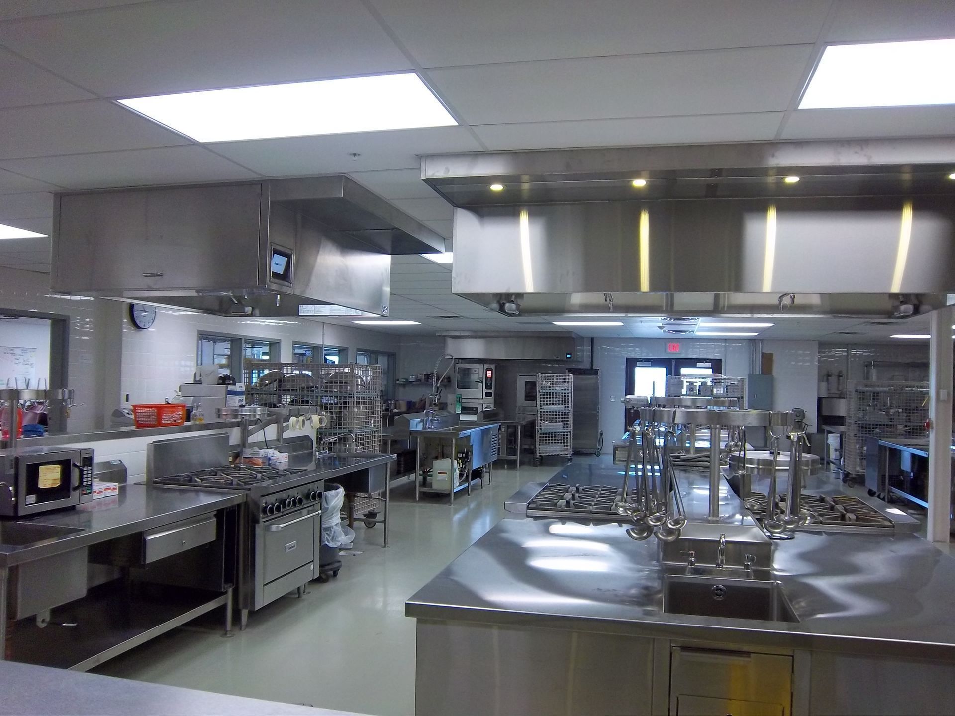 A clean, commercial kitchen with stainless steel countertops, industrial appliances, and large overhead ventilation hoods.
