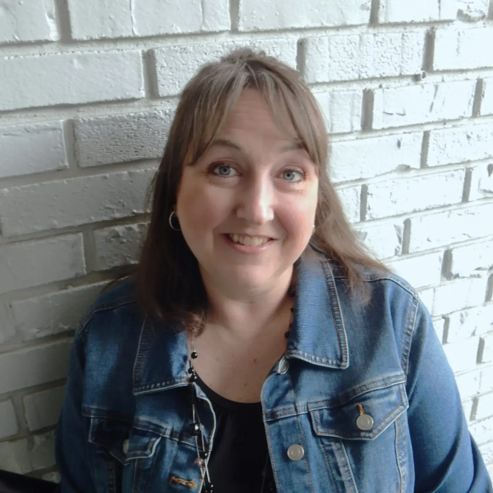 A smiling person wearing a blue denim jacket over a black top, positioned in front of a white brick wall.
