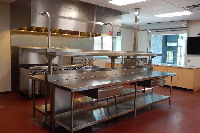A stainless steel prep table with shelves sits in a commercial kitchen with a hood vent and reddish-brown flooring.