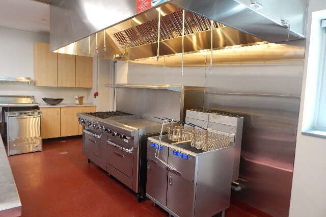Commercial kitchen with stainless steel stoves and a deep fryer under a metal ventilation hood on a red floor.