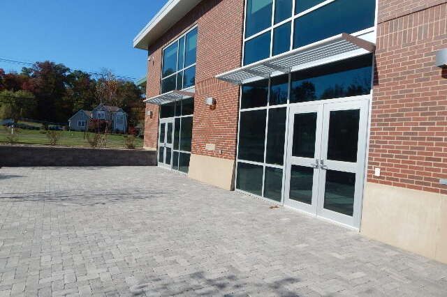 A modern brick building entrance with large glass windows, double glass doors, and a paved stone patio area.