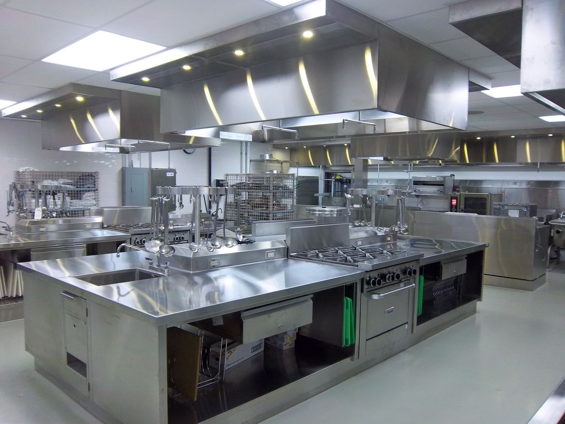 Commercial kitchen with stainless steel workstations, multiple ovens, a large stove, and overhead ventilation hoods.
