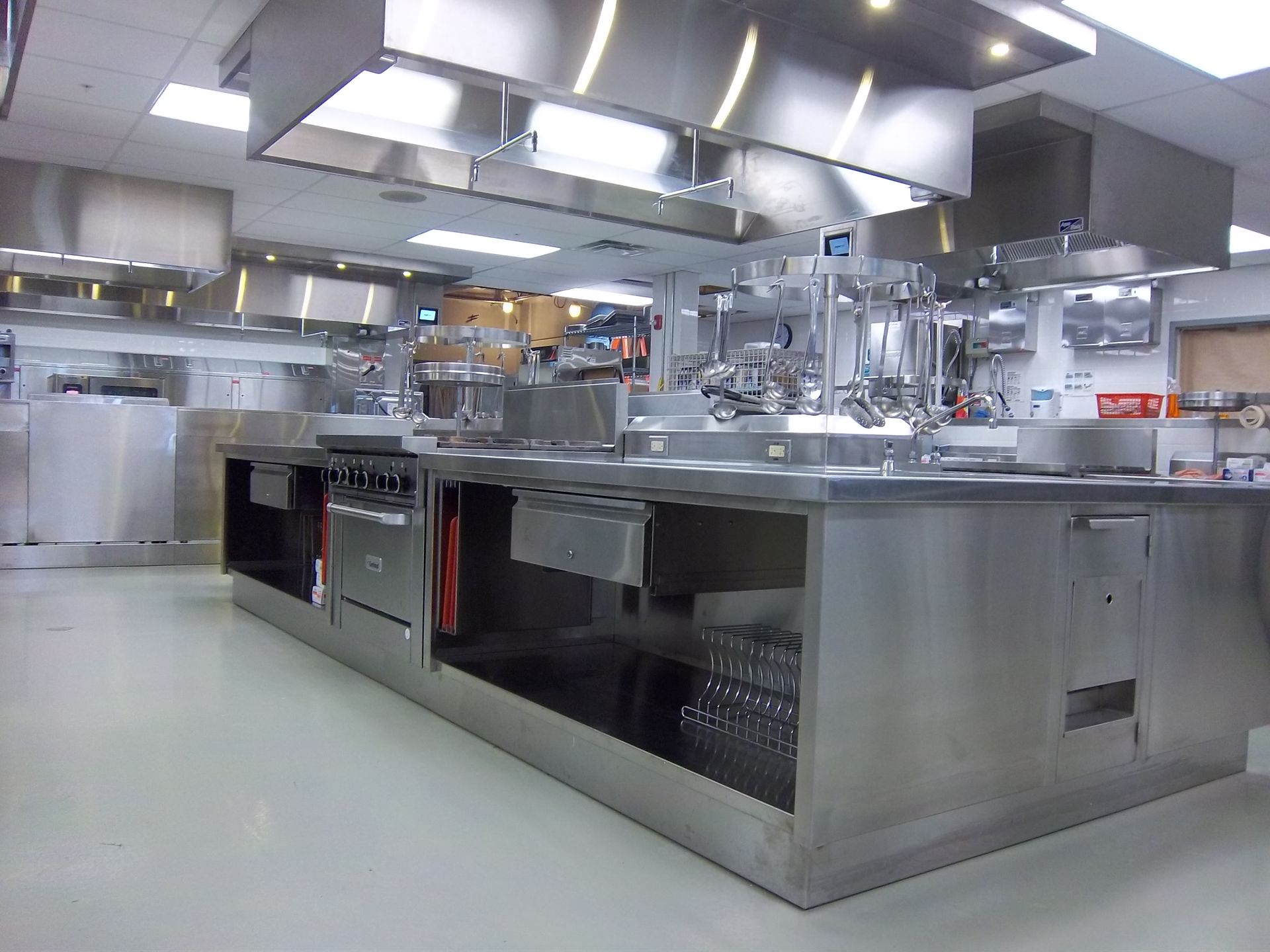 Stainless steel commercial kitchen with a central prep island, multiple ranges, and industrial exhaust hoods overhead.