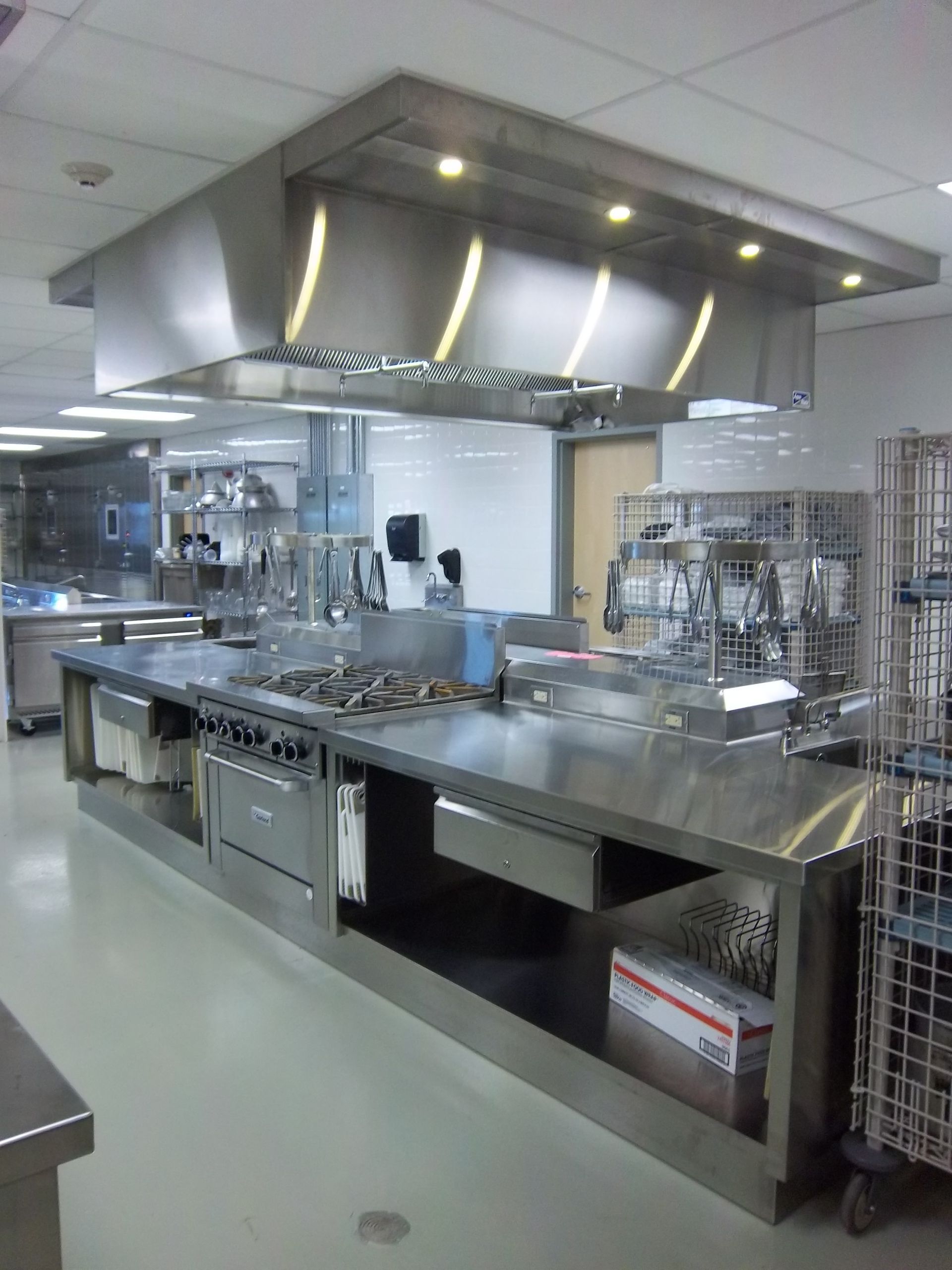 A stainless steel commercial kitchen island with a gas range and an overhead exhaust hood in a tiled room.