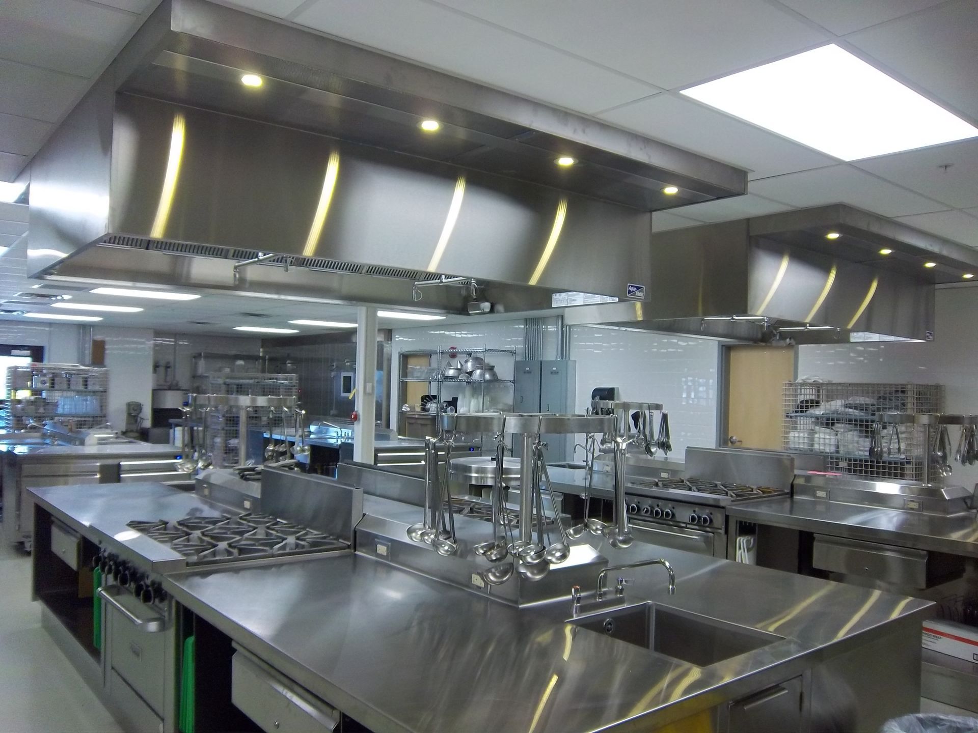 A professional stainless steel commercial kitchen featuring multiple cooking stations with overhead ventilation hoods.