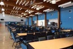 A large, empty cafeteria with rows of rectangular tables and chairs under a high wooden ceiling with modern lighting.