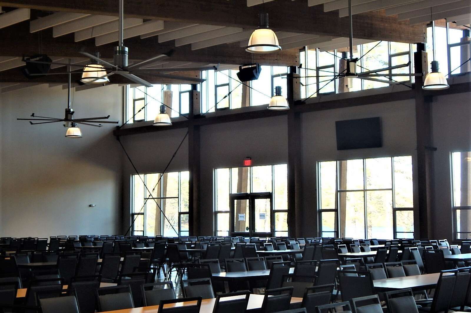 A large, empty dining hall with rows of tables and chairs, tall windows, exposed wooden ceiling beams, and pendant lights.